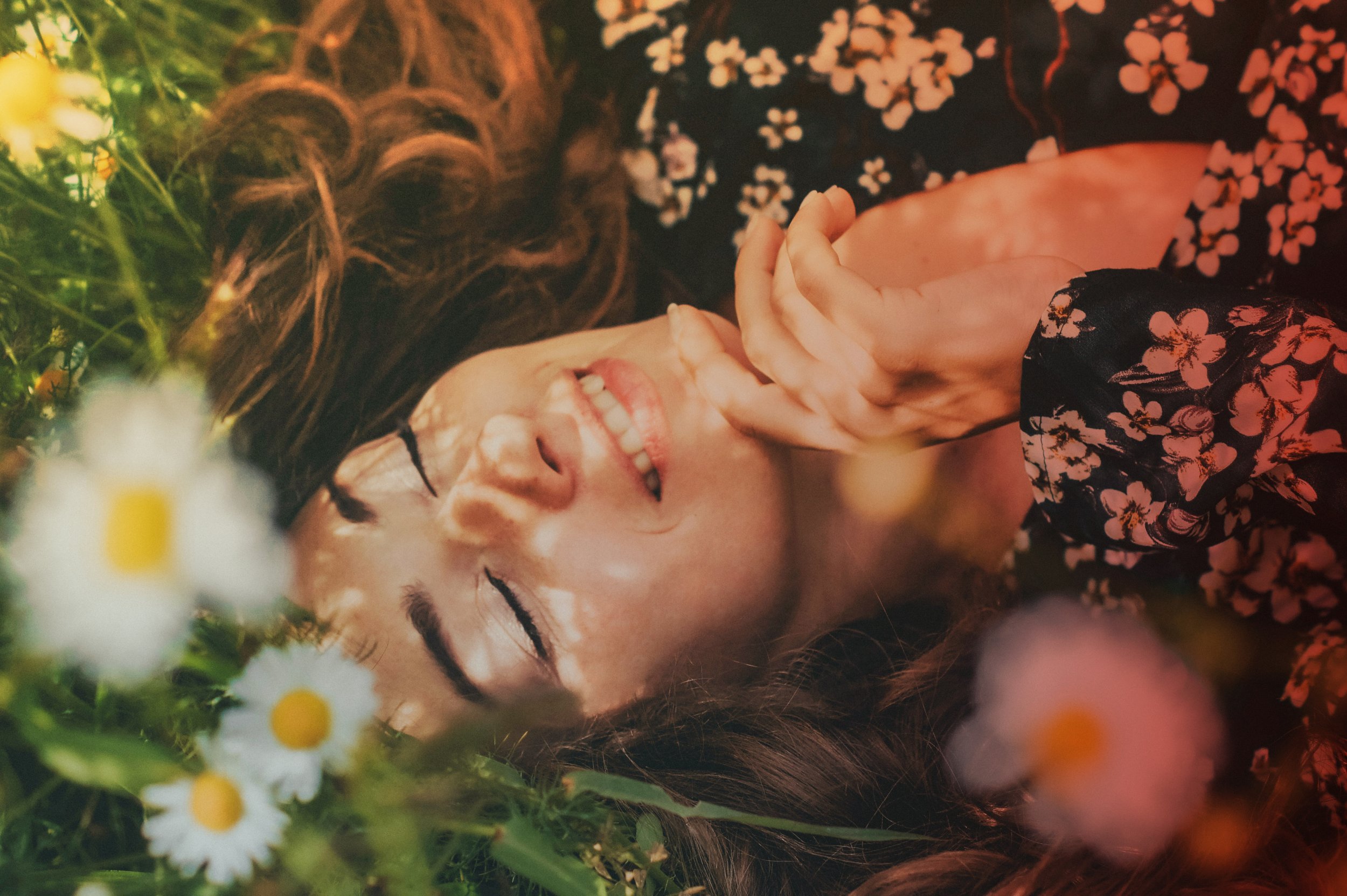 Femme souriante avec yeux fermés, allongée dans des fleurs sauvages, portant une robe à motifs floraux, main près du visage, cheveux bruns, ambiance chaleureuse et naturelle.