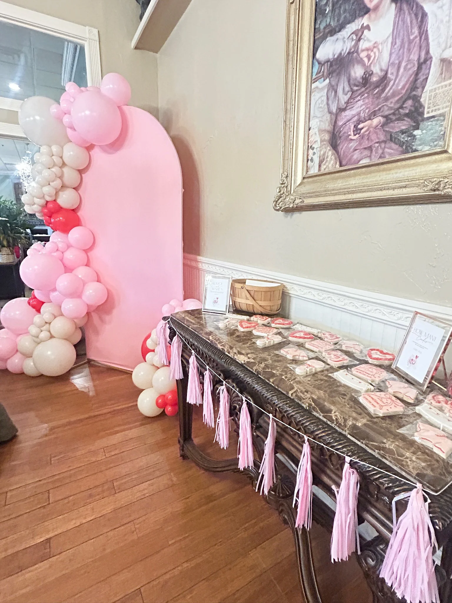 Decorative pink balloon arch with white and red balloons next to a pink backdrop, set up for a celebration or party. A marble-top table decorated with pink tassels and various pink and white treat cookies is in the foreground, and a framed painting h
