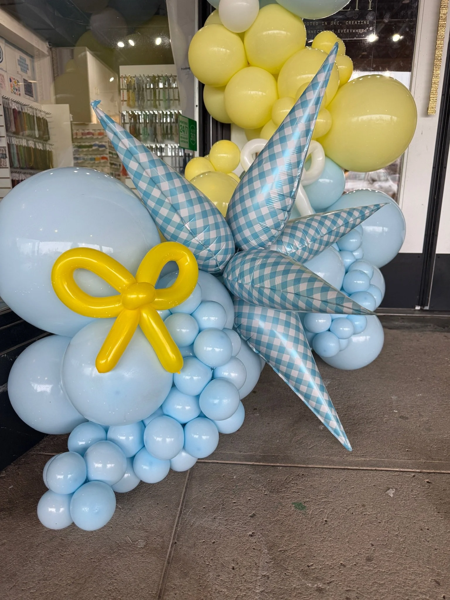 Balloon display featuring light blue, yellow, and white balloons with a blue and white checked pattern, arranged in a cluster outside a store.