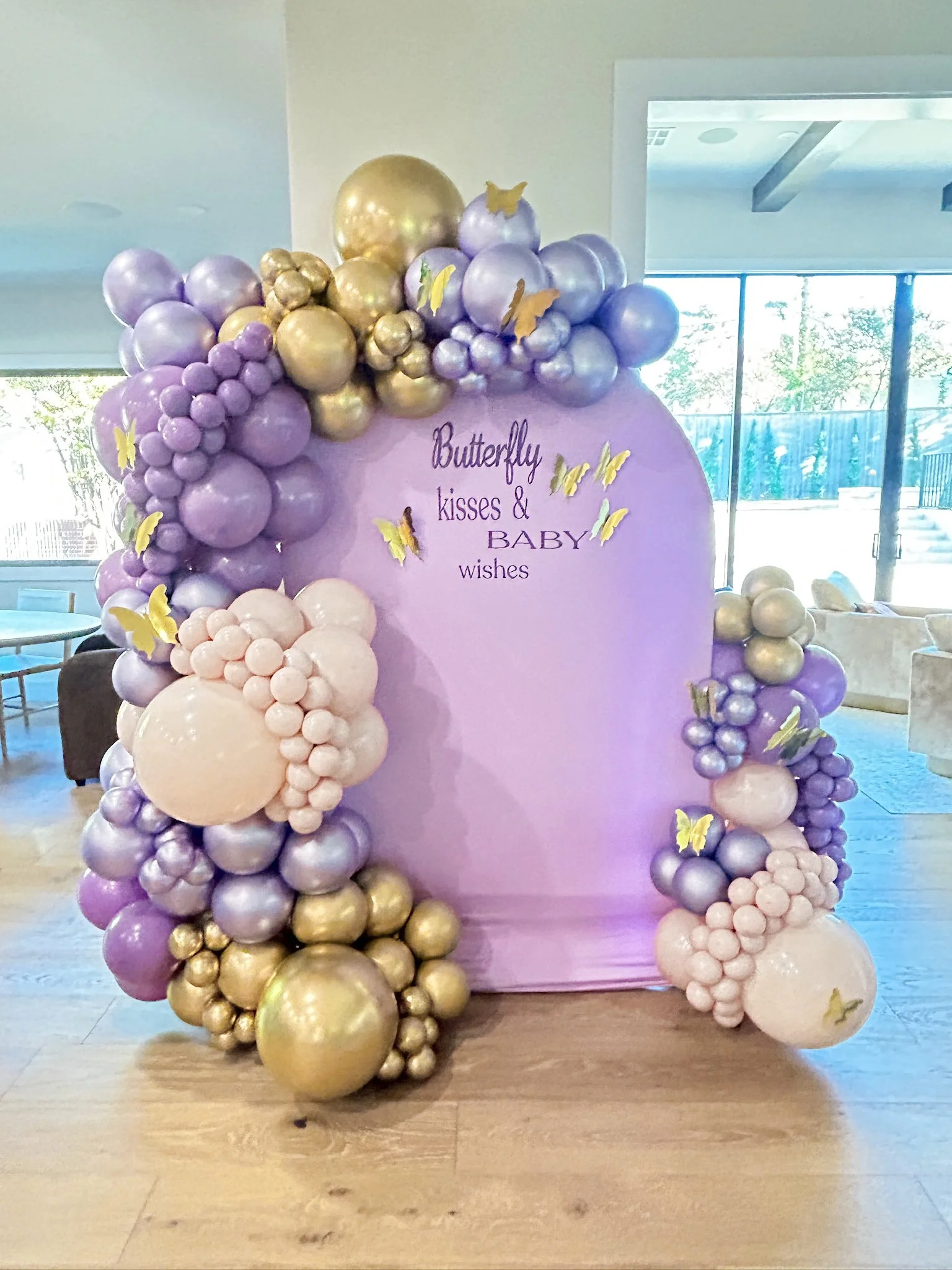 Decorative backdrop with purple, pink, and gold balloons surrounding a purple circle, featuring yellow butterfly decals and the text 'Butterfly kisses & BABY wishes' in the center.