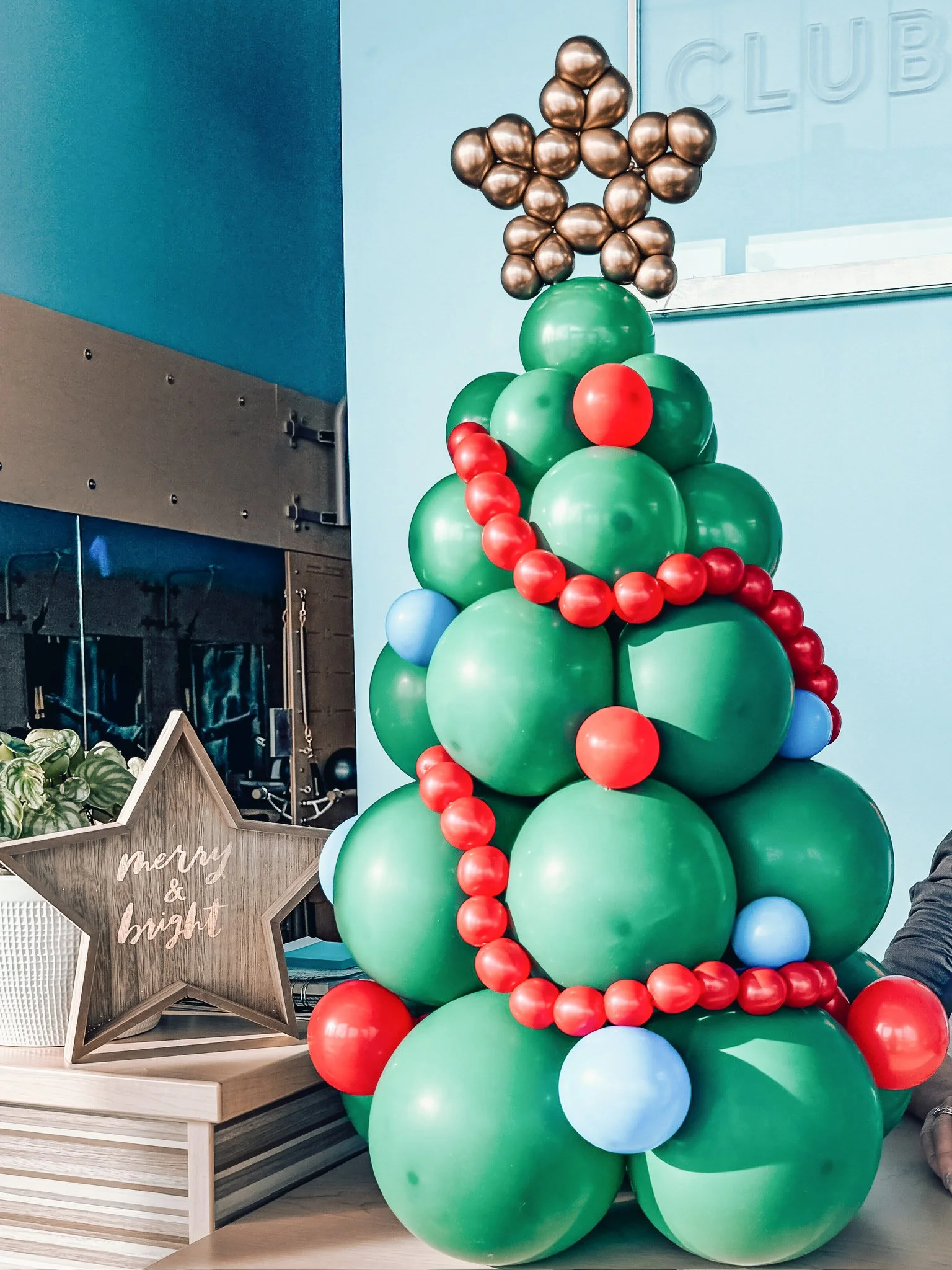 Balloon Christmas tree decorated with red, blue, and bronze balloons, with a star-shaped sign that says "merry & bright" in the background.