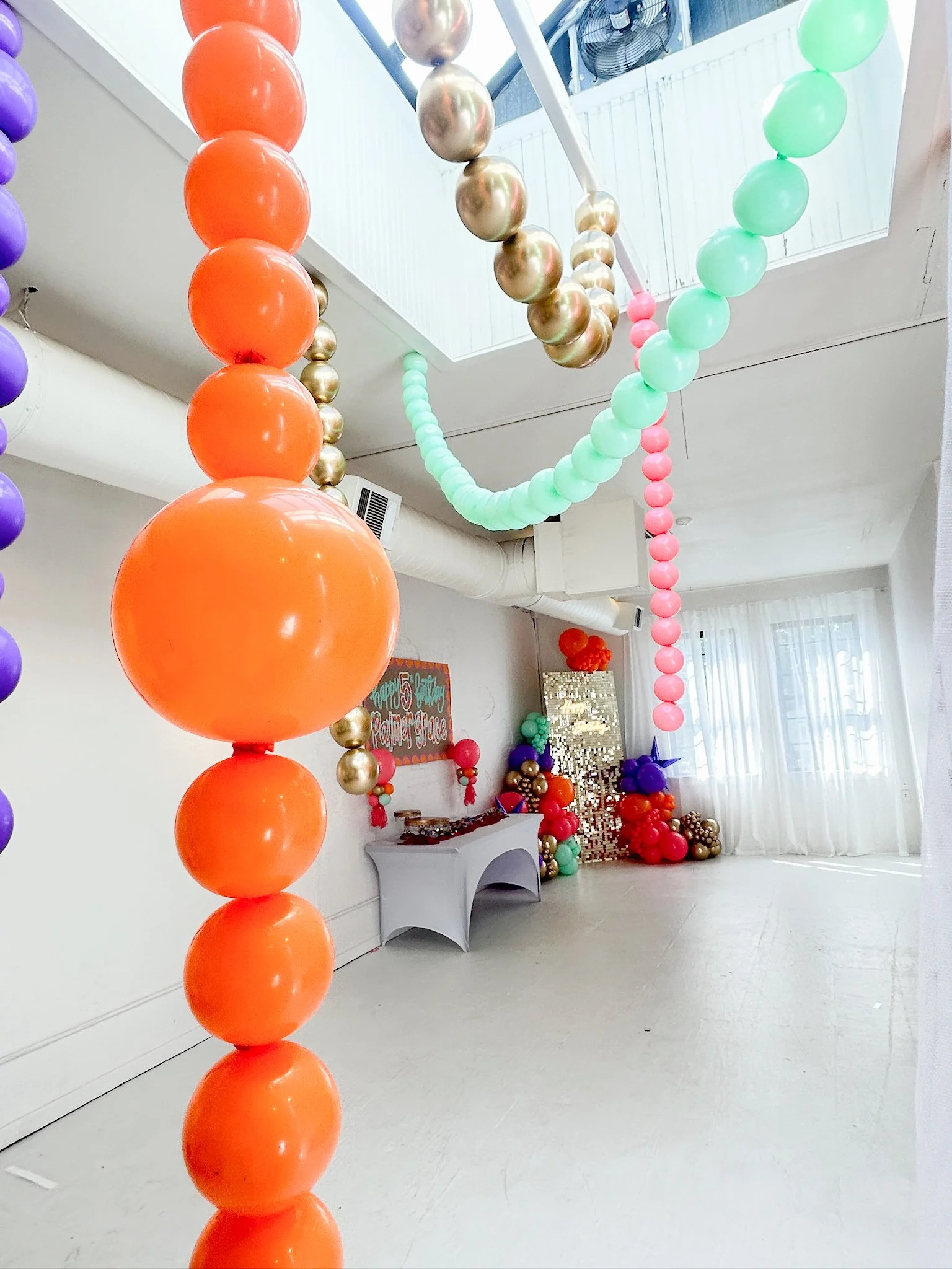 Colorful balloon decorations hanging from the ceiling, including large orange, gold, mint, pink, and purple balloons, with a table and a decorative backdrop in the background for a celebration or party.