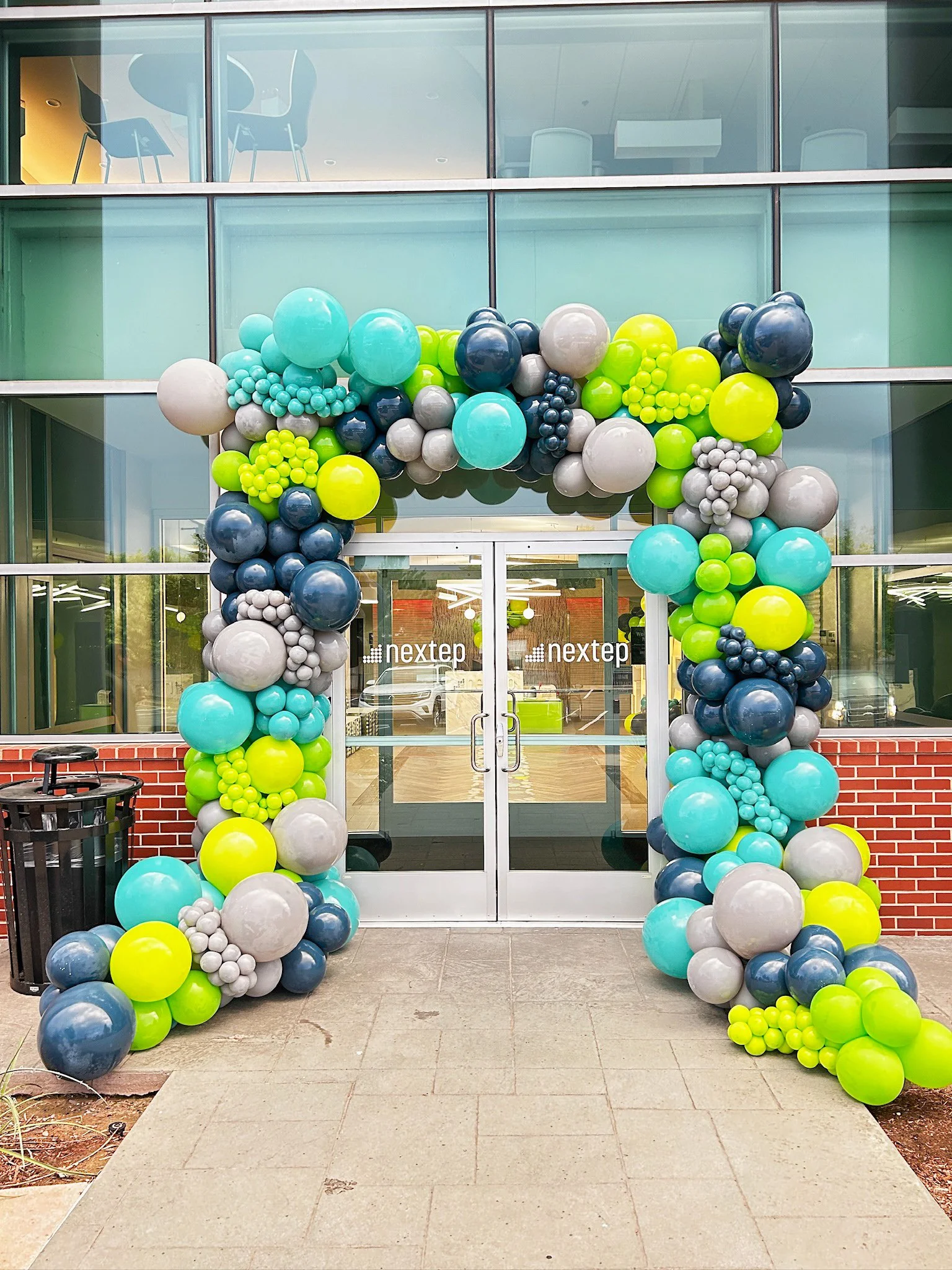 Colorful balloon arch outside a glass building entrance with doors marked 'Nextep'.
