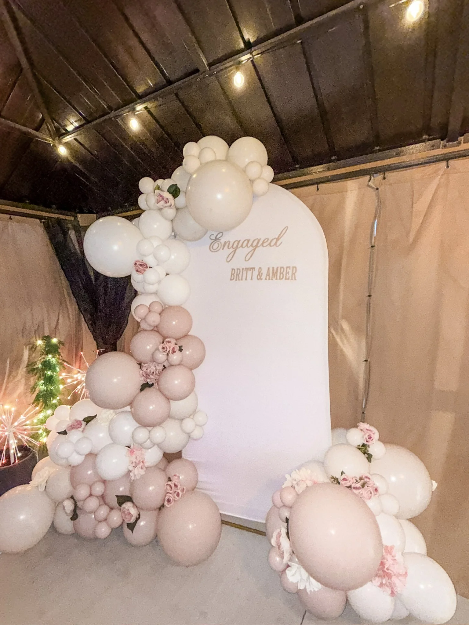 Decorative backdrop with white and blush pink balloons, flowers, and greenery for an engagement celebration. A white sign with gold lettering says "Engaged" and the names Britt and Amber.