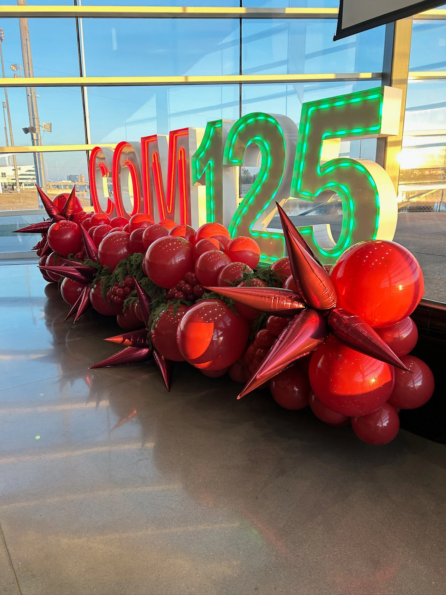 Decorative display with large illuminated "COM 125" sign in neon green and red, surrounded by red balloons and star-shaped decorations, inside a building with large glass windows showing a blue sky.