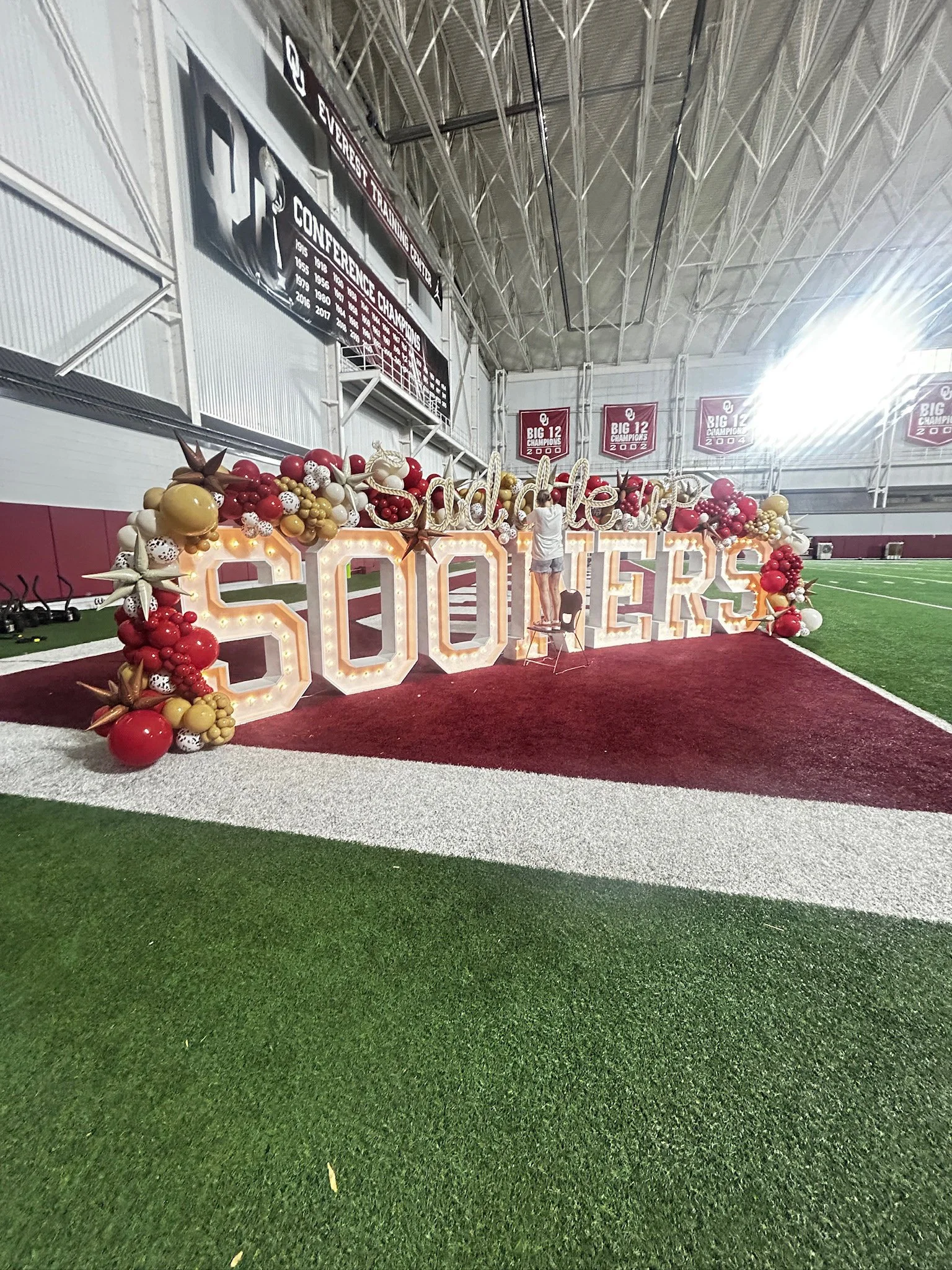 Decorative sign with large illuminated letters spelling 'Sisters' inside a sports stadium with banners and a green artificial turf field.