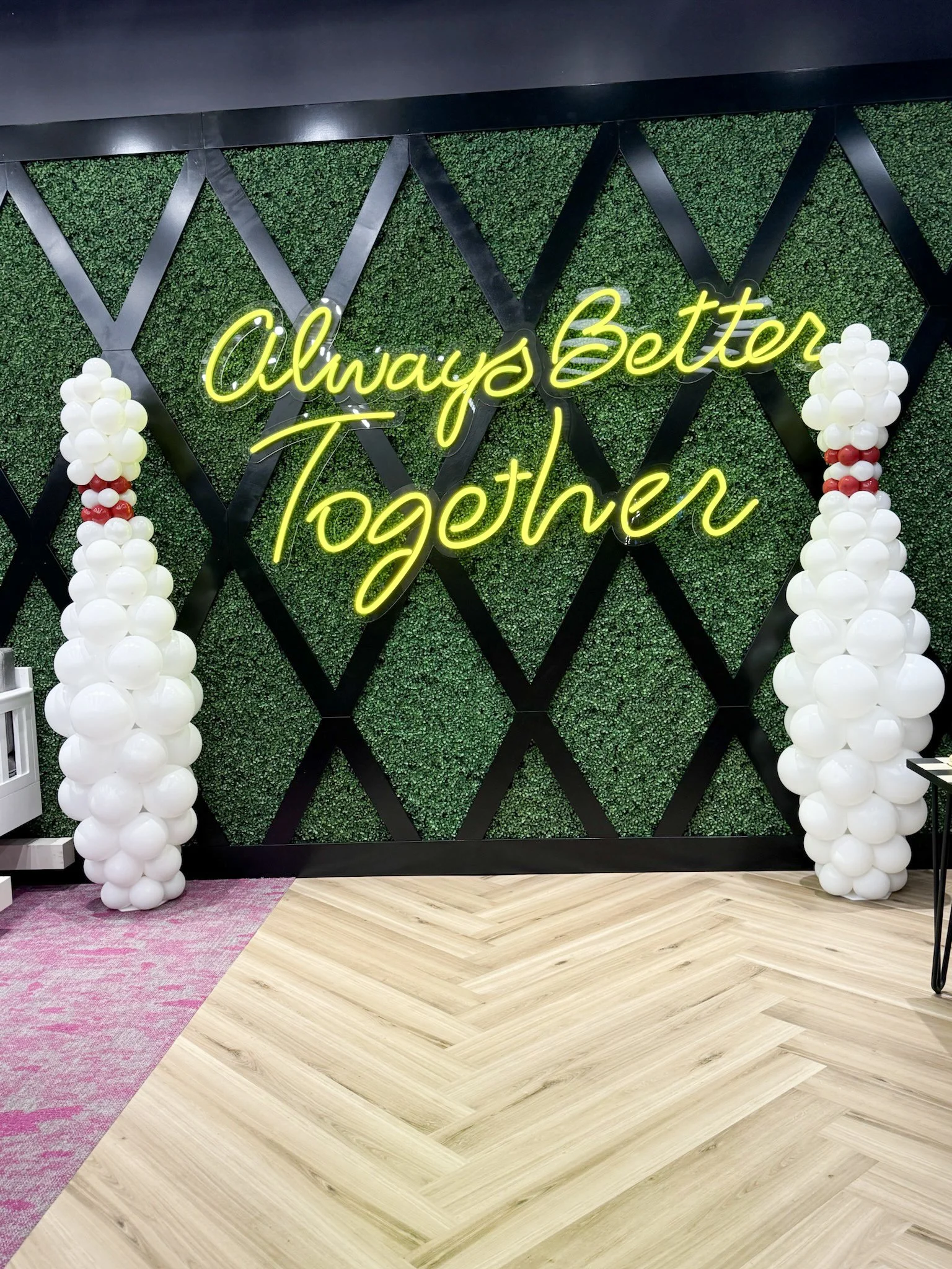 Decorative display with a neon sign that says 'Always Better Together,' flanked by two columns of white balloons with red accents, on a wall with green foliage and a black geometric frame.
