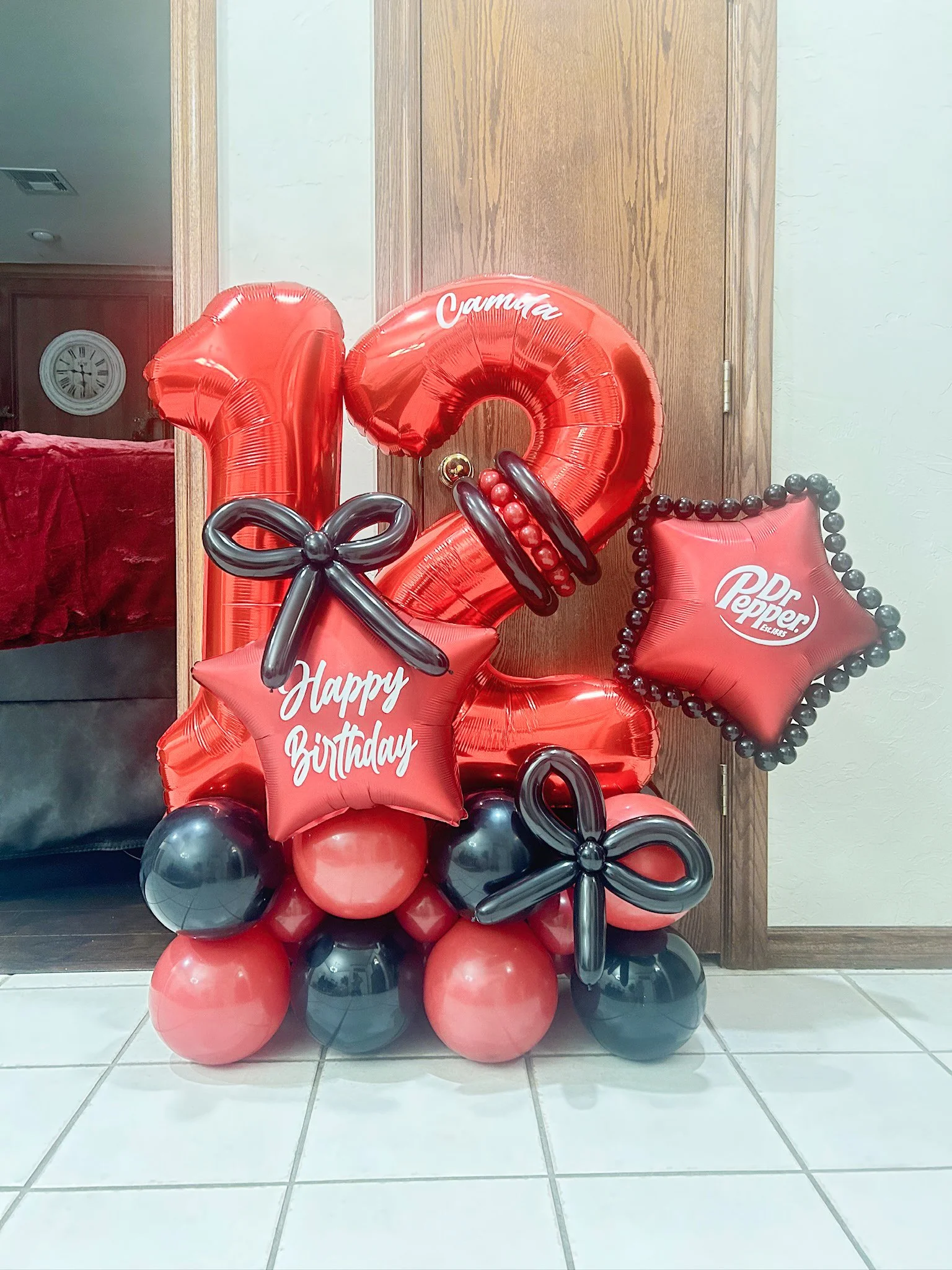 Balloon arrangement with red and black balloons celebrating a 12th birthday, featuring large number balloons '1' and '2', and star-shaped balloons with messages 'Happy Birthday' and logos of Dr. Pepper and Camila.
