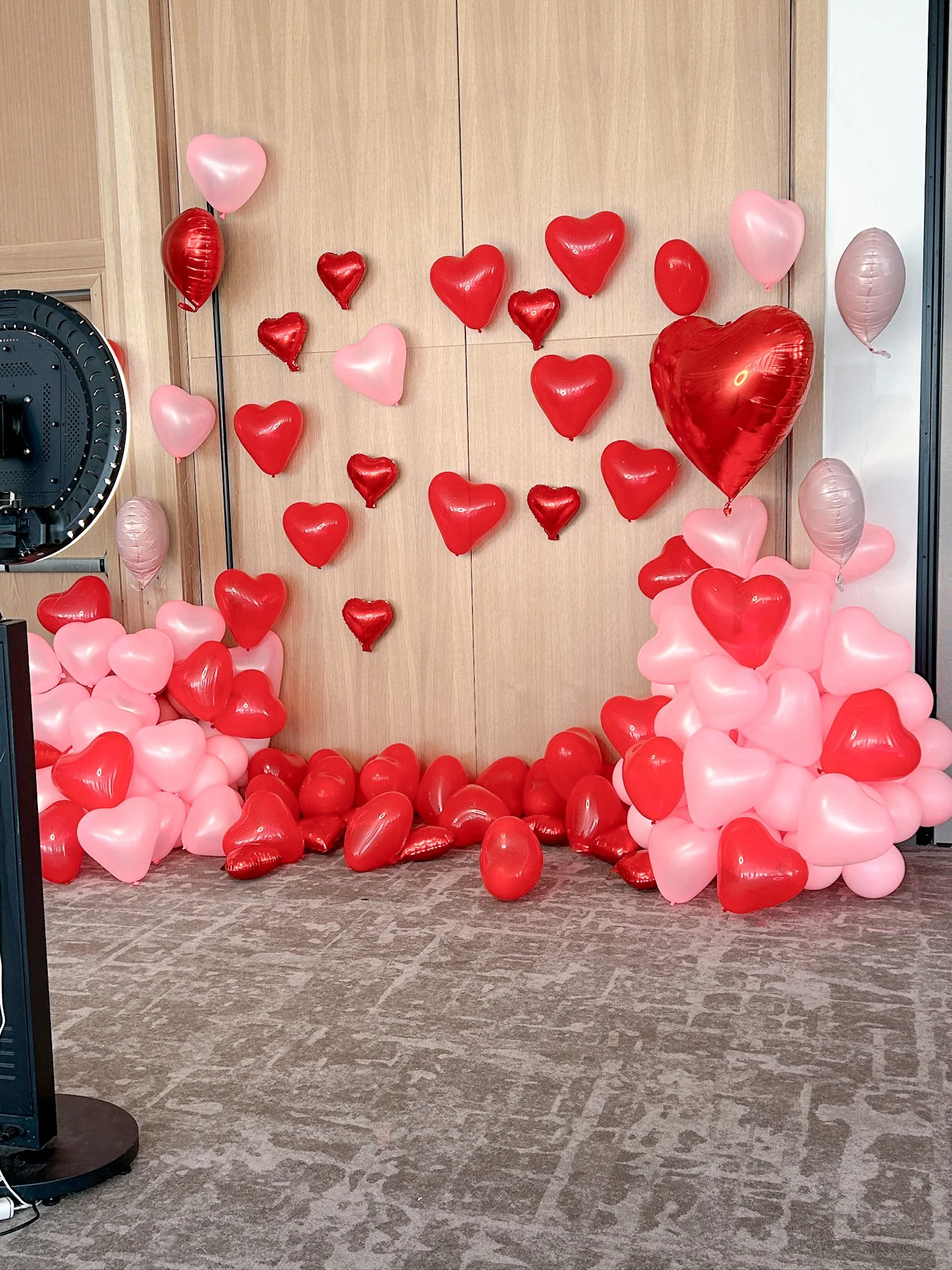 Decorative backdrop with red and pink heart-shaped balloons, some floating and some on the floor, arranged against a wooden wall for a celebration or romantic event.
