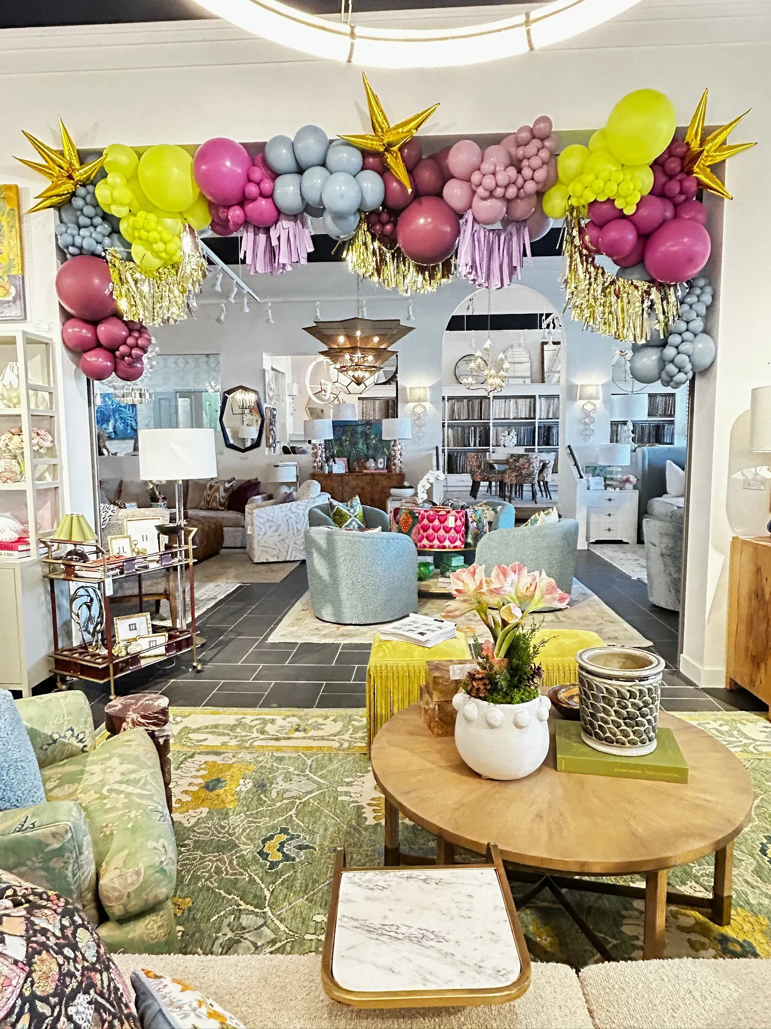 Decorative balloon and star garland in pastel pink, yellow, blue, and gold colors hanging above a bright living room with assorted furniture and plants.