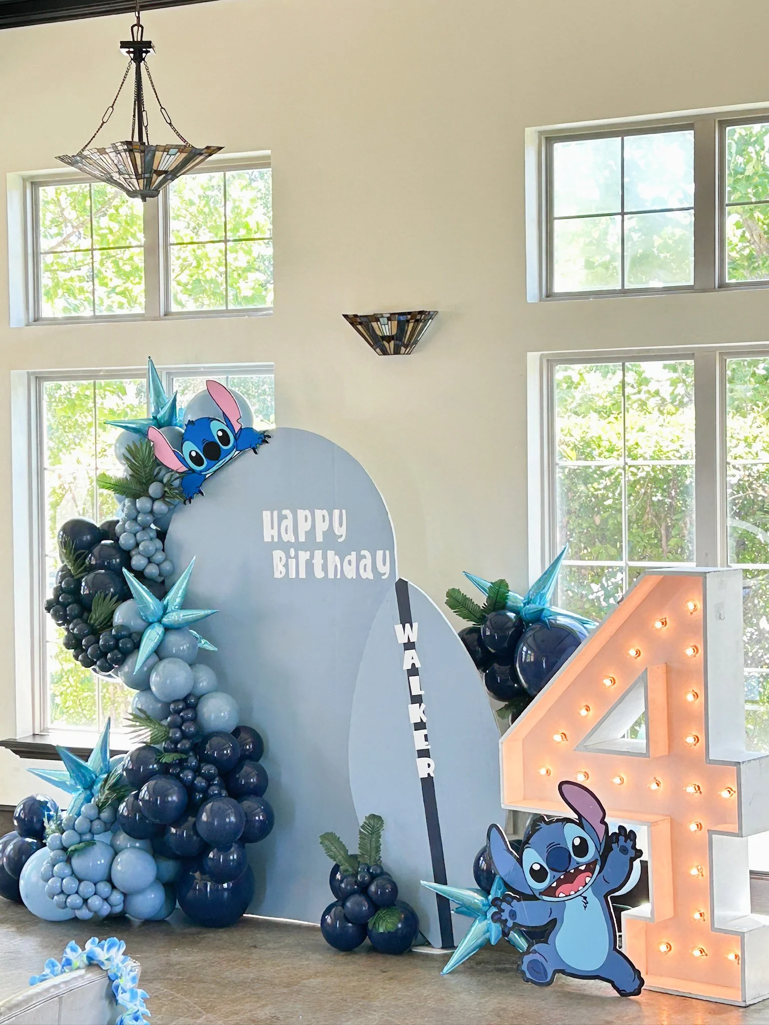 Colorful birthday decoration featuring a 'Lilo & Stitch' theme with balloon arrangements, a large illuminated number four, and a backdrop with the message 'Happy Birthday'.