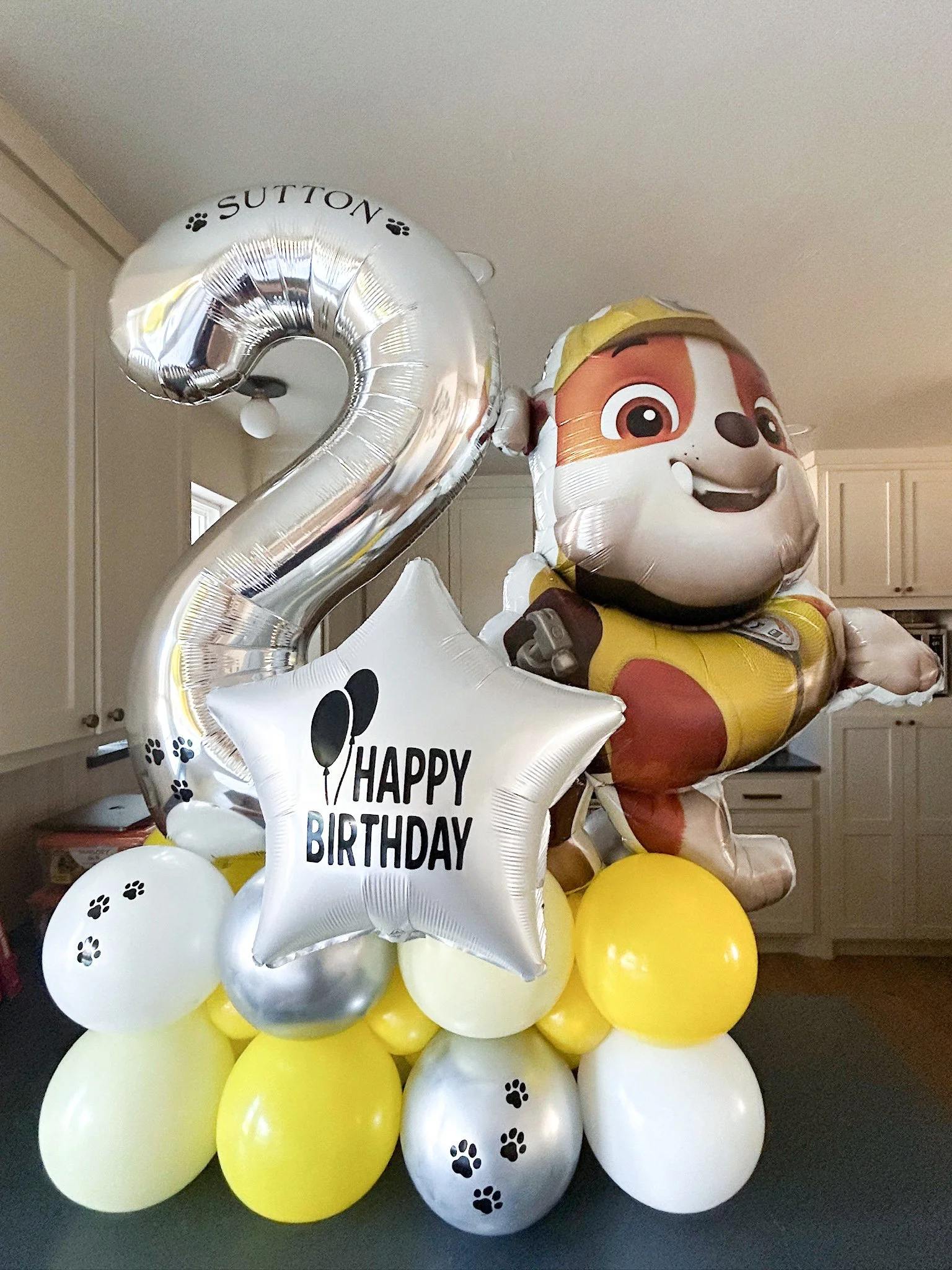 Balloon arrangement with a large number two, a star-shaped balloon with 'Happy Birthday,' and a puppy character balloon, all in yellow, white, and silver colors.