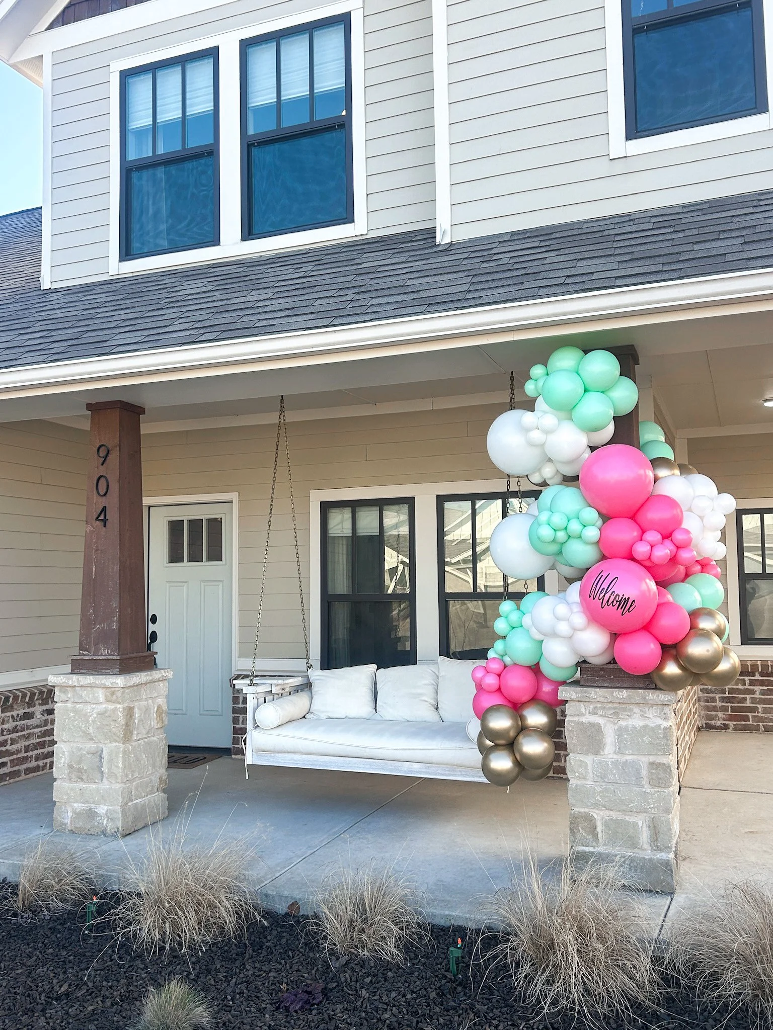 Front porch of a house decorated with a balloon arch and a swing with white cushions, welcoming guests.