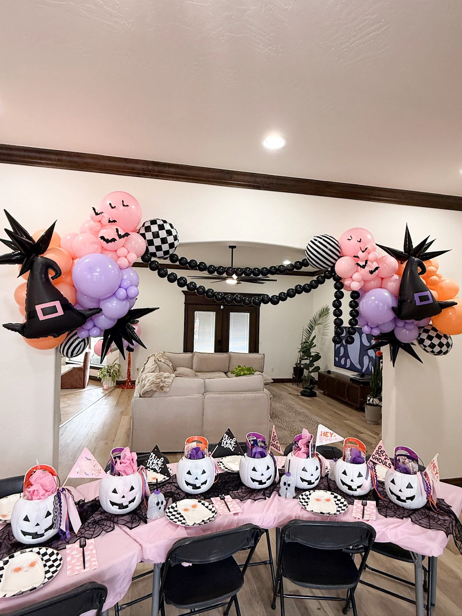 A Halloween-themed party setup in a living room with a table decorated for kids, featuring white pumpkin-themed buckets with spooky faces, black spider web tablecloth, pink and black themed decorations, and balloon arrangements with witches' hats, st