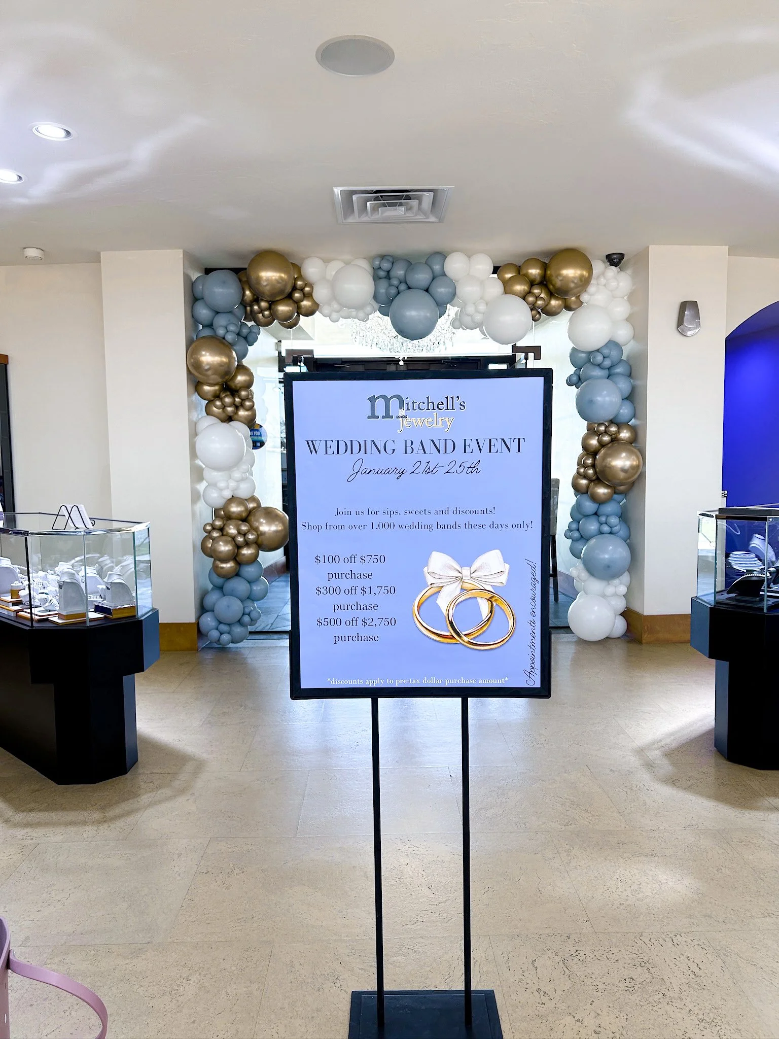 Sign for Mitchell's Jewelry Wedding Band Event from January 21st to 25th, featuring promotional discounts, with balloon decorations in white, gold, and light blue surrounding the entrance.