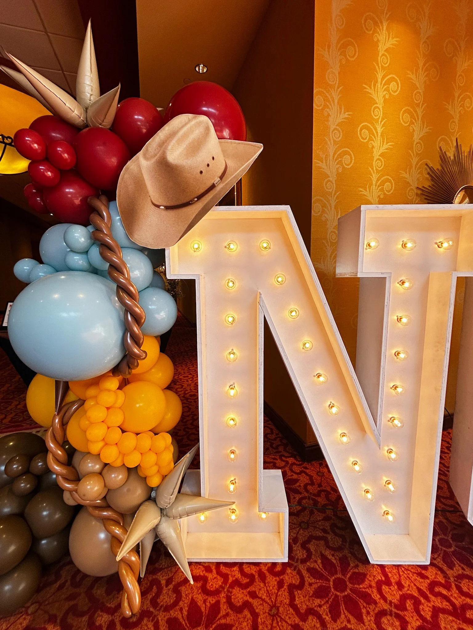 A large decorative letter 'N' with lights inside, surrounded by colorful balloons and cowboy-themed decorations like a cowboy hat, star, and twisted balloon ropes, for a festive event.