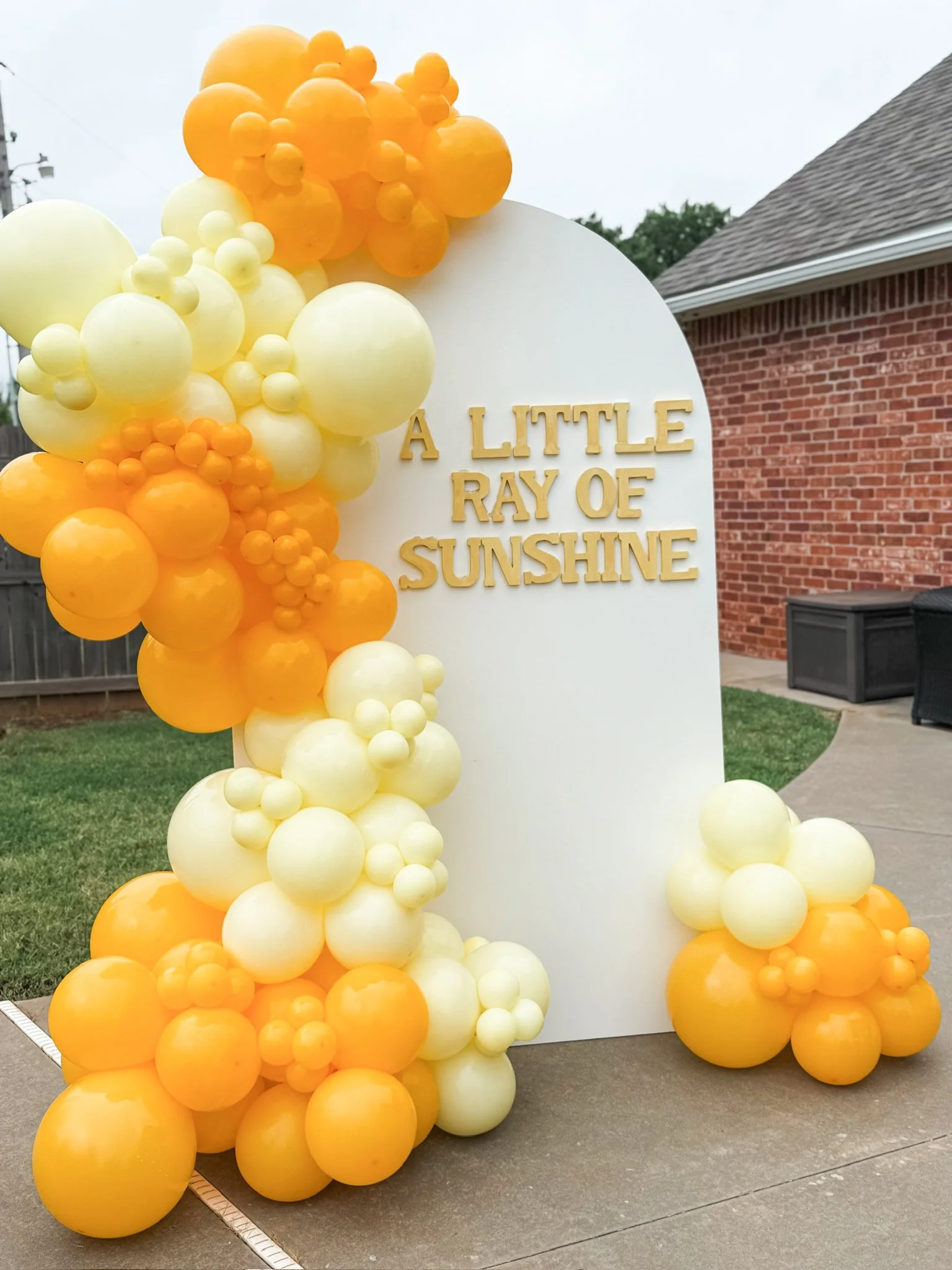 Decorative outdoor display with white, yellow, and orange balloons arranged around a white curved board that has the text 'A LITTLE RAY OF SUNSHINE' in gold letters.