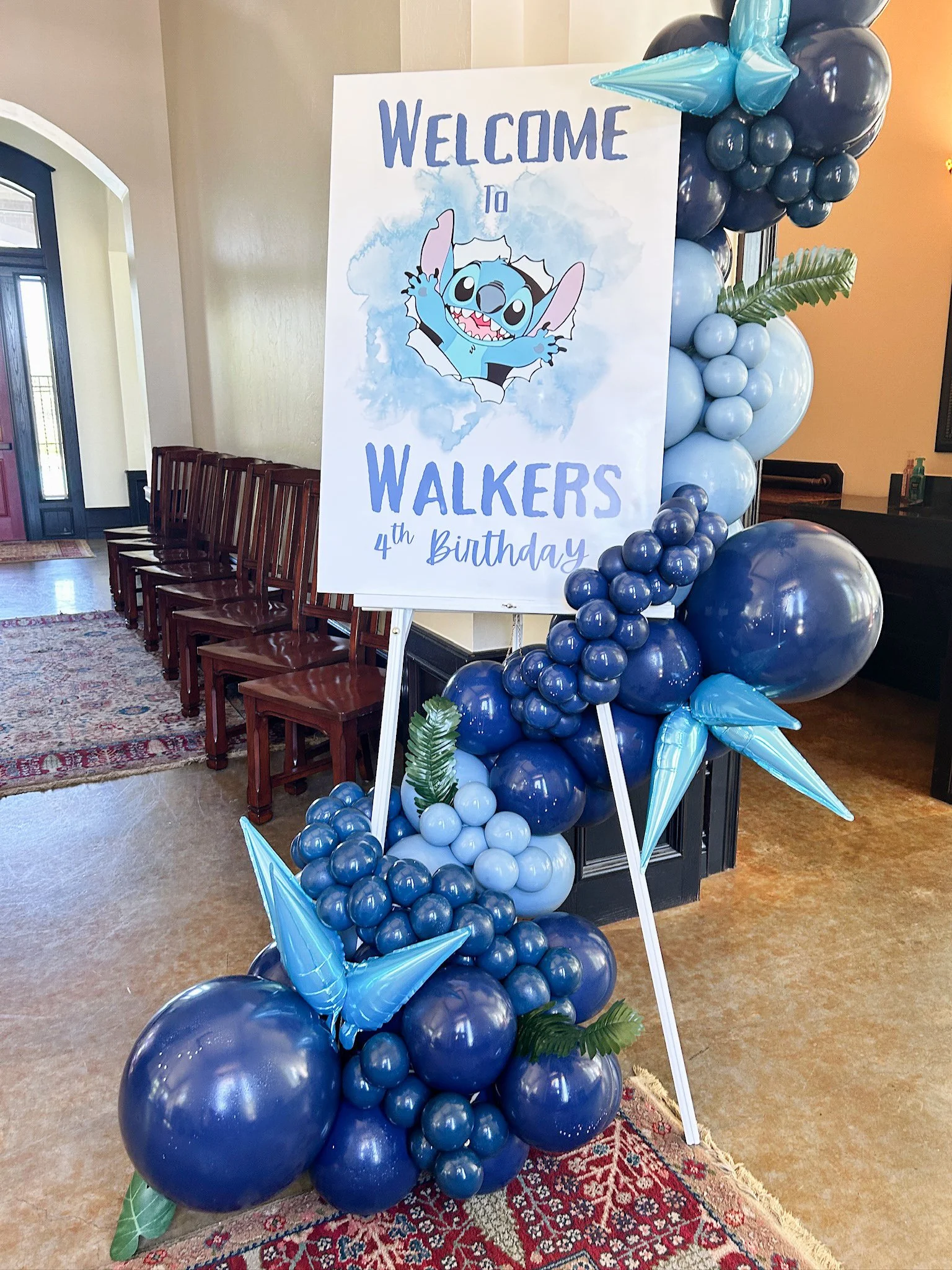 Decorative welcome sign for 'Stitch' character's 4th birthday, surrounded by blue balloons and tropical leaves.