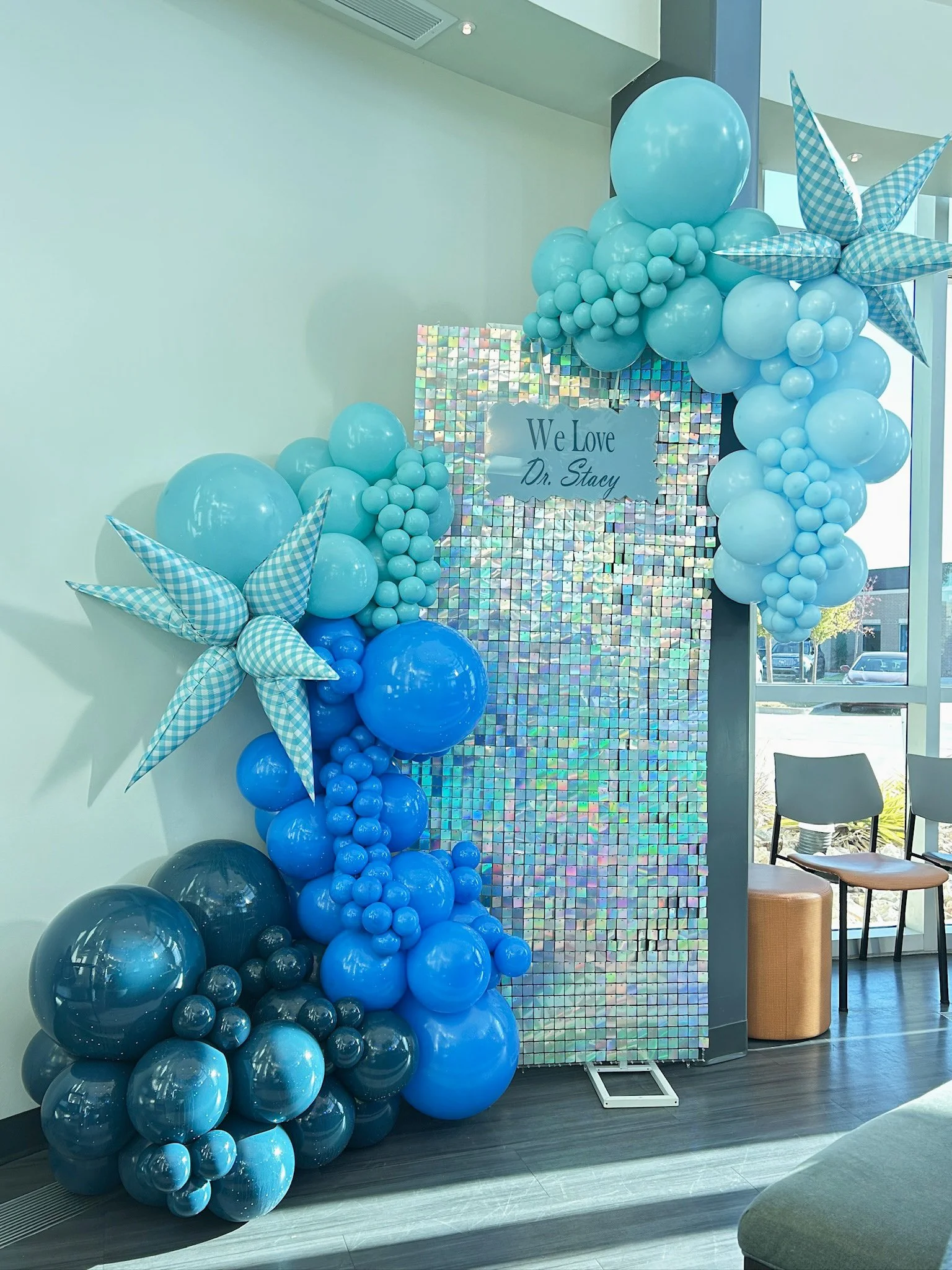 Decorative balloon arch with blue balloons, paper flowers, and a mirrored backdrop with a sign that says "We Love Dr. Stacy" in a bright room with chairs and a window.