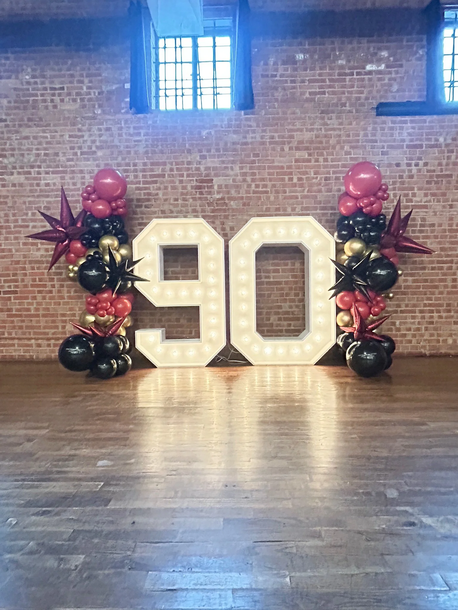 Large illuminated number '90' decorated with balloons in black, pink, and gold, arranged on a wooden floor against a brick wall with two windows.