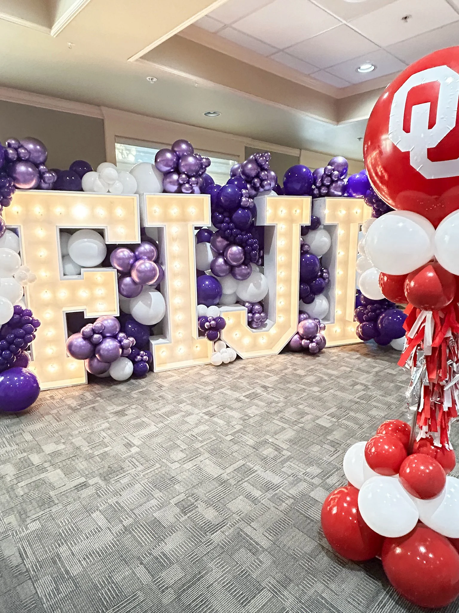 Large decorative marquee letters spelling 'FUT' outlined with warm lights, surrounded by clusters of purple, white, and silver balloons, with additional red and white balloons on the right side, set in an indoor venue with carpeted floor and ceiling 