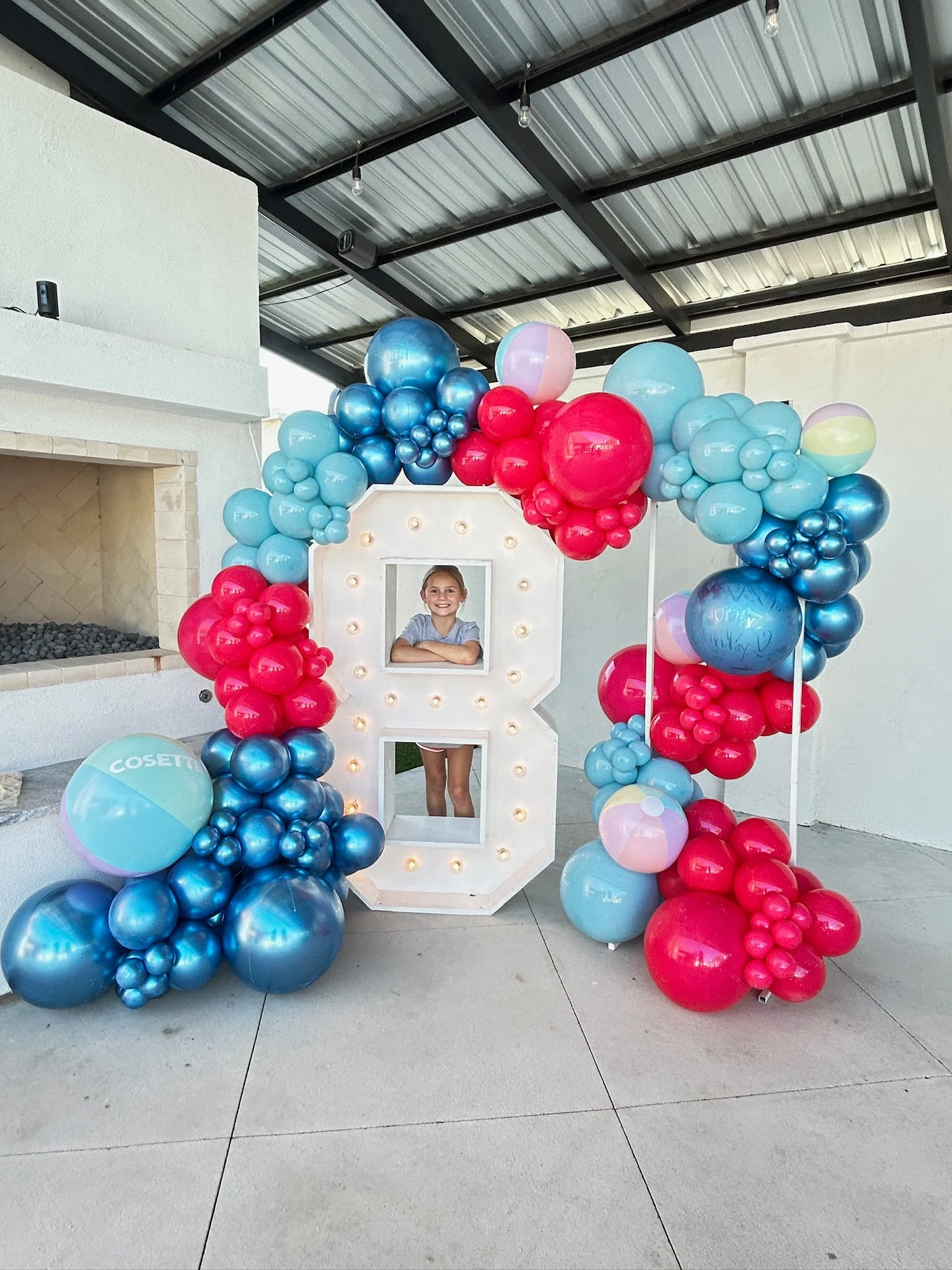 A girl standing behind a large decorated letter 'B' with light bulbs, surrounded by colorful balloons, at an outdoor party or celebration.