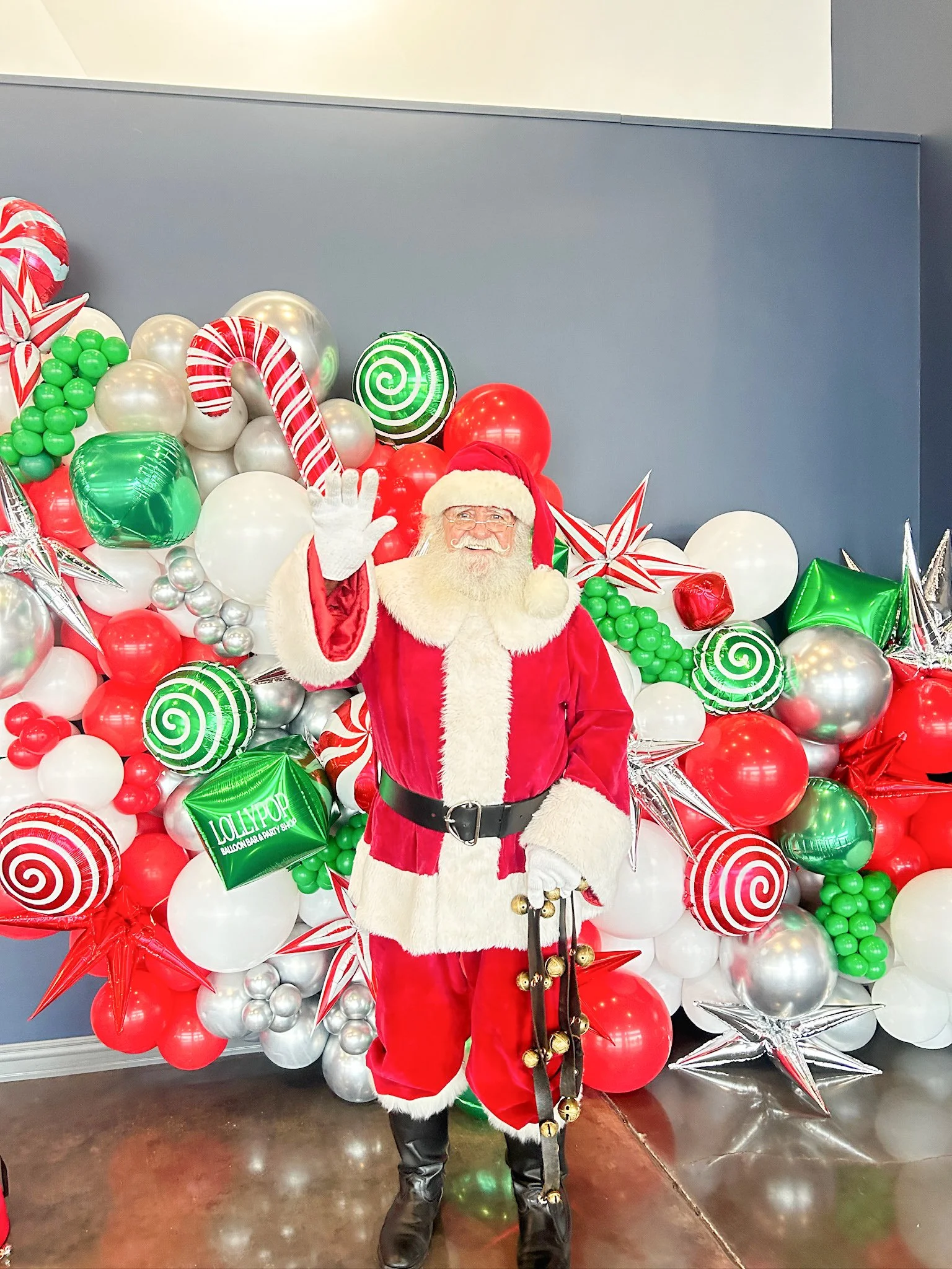Santa Claus in a red suit and hat, waving and standing in front of a festive Christmas balloon display with red, white, green, and silver balloons, including candy canes and star-shaped balloons.