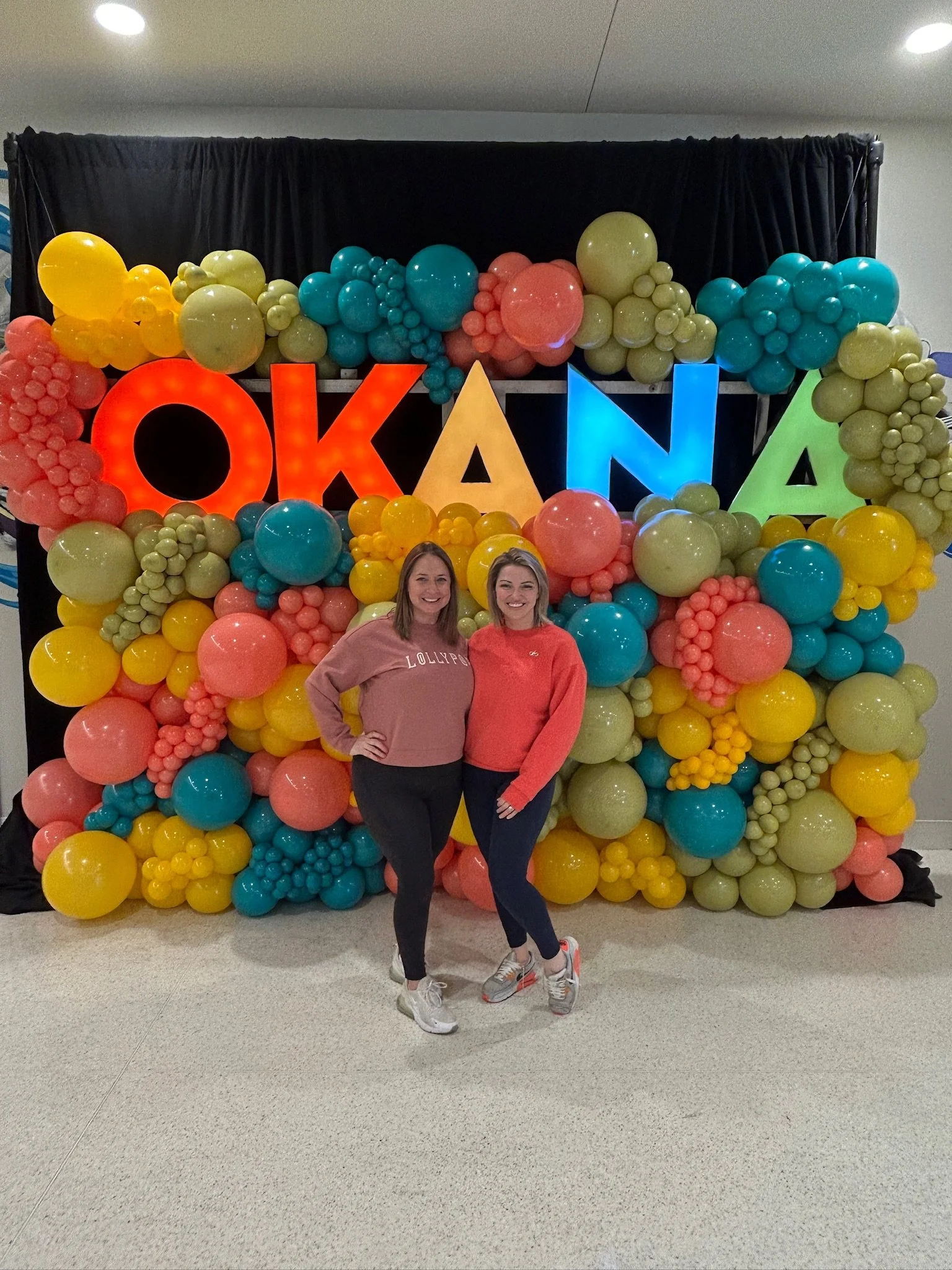 Two women standing in front of a colorful balloon display with the word 'OKLAHOMA' in illuminated letters.