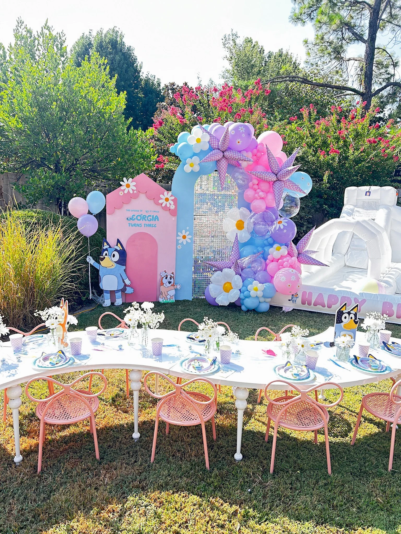 Colorful birthday party setup with pink, purple, and blue balloons, a balloon arch, cartoon character cutouts, a long white table with pink chairs, flower arrangements, and a decorated backdrop featuring a cartoon dog and a dog house indicating a bir