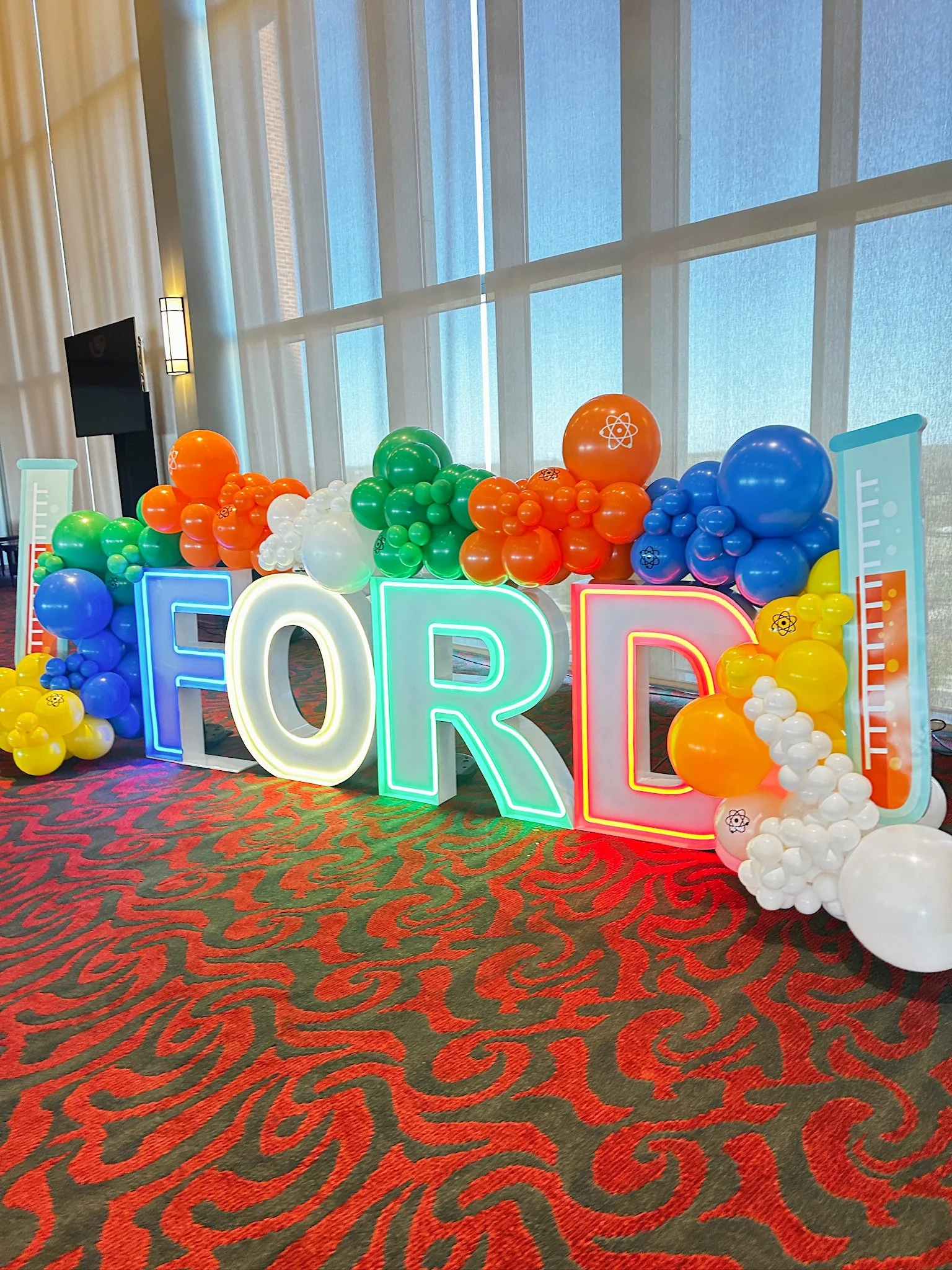 Colorful balloon arrangement with large illuminated letters spelling 'FOR' and scientific-themed decorations at an event venue.