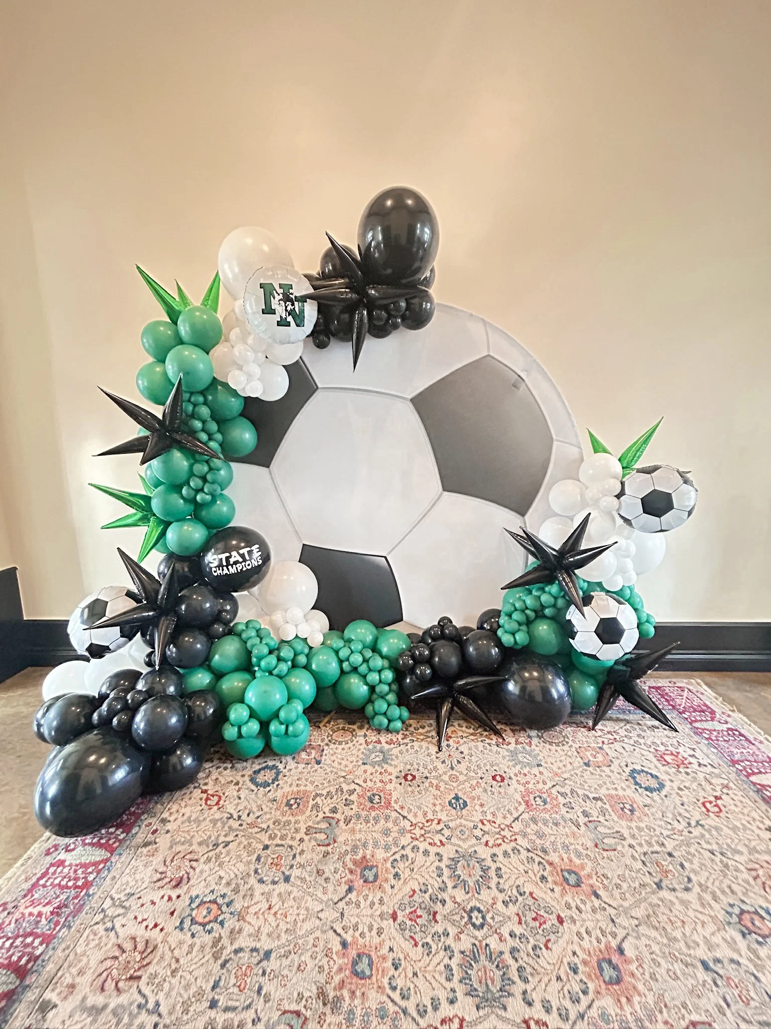 Balloon display with a soccer theme, including a large soccer ball backdrop, black, white, green, and teal balloons, some shaped like soccer balls and trees, with star-shaped black balloons and a balloon that says 'State Champions'.
