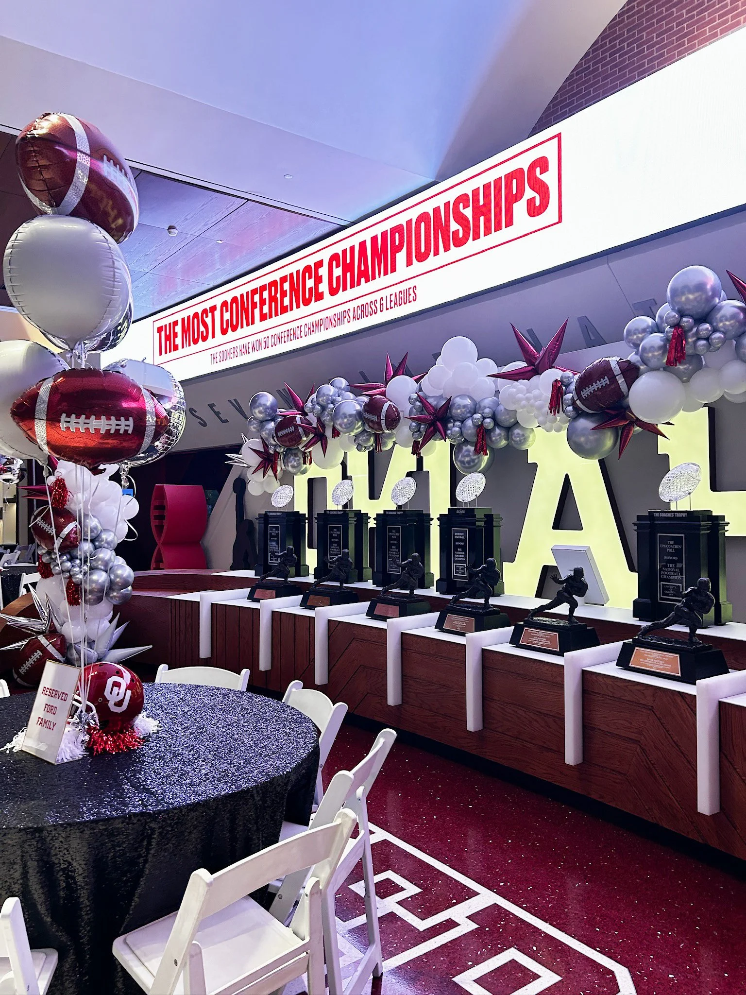 Decorated indoor space with balloons, trophies, and a sign celebrating the conference championships in American football.