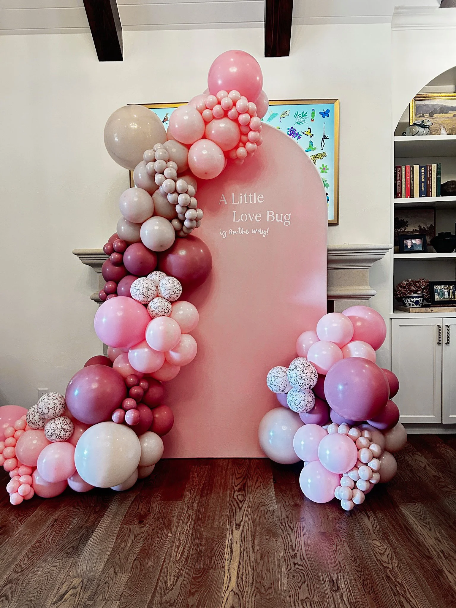 Decorative balloon arch and backdrop with pink, white, and maroon balloons surrounding a pink board that reads 'A Little Love Bug is on the way!'