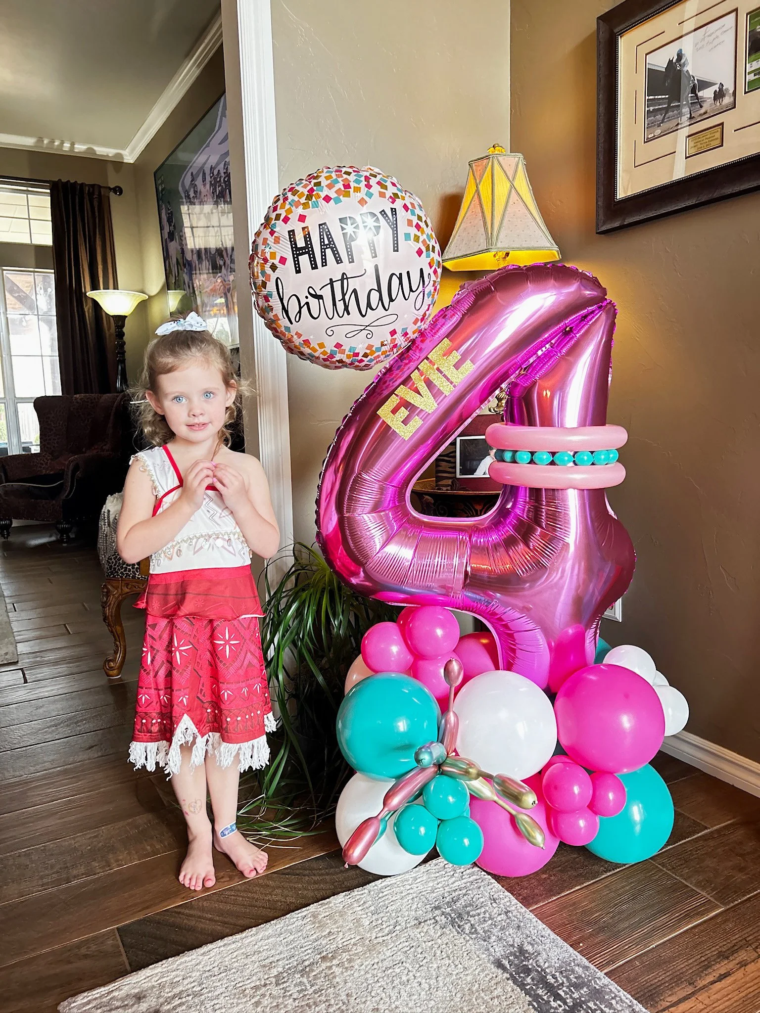 A young girl in a white top with red accents and a pink skirt with white fringe stands next to a large pink number four balloon with the name 'EVE' and a crown on it. The balloons are part of a birthday decoration, including a string of balloons at t