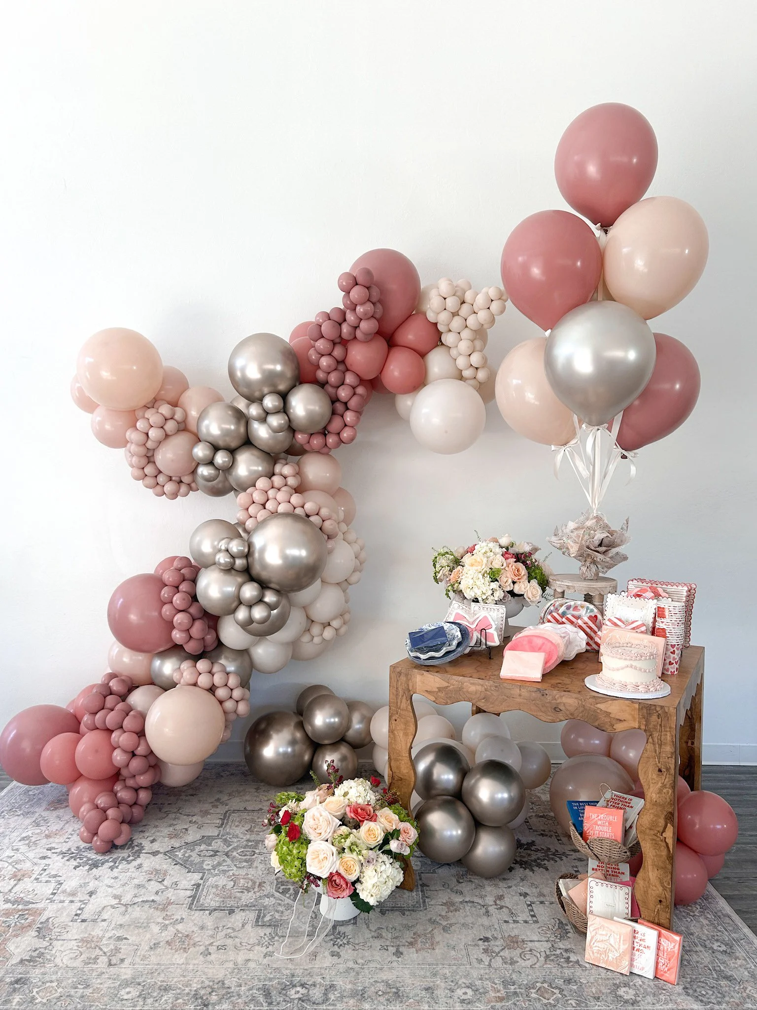 Decorative arrangement featuring pink, white, and silver balloons, a bouquet of flowers, gifts, and a small cake displayed on a wooden table against a white wall.
