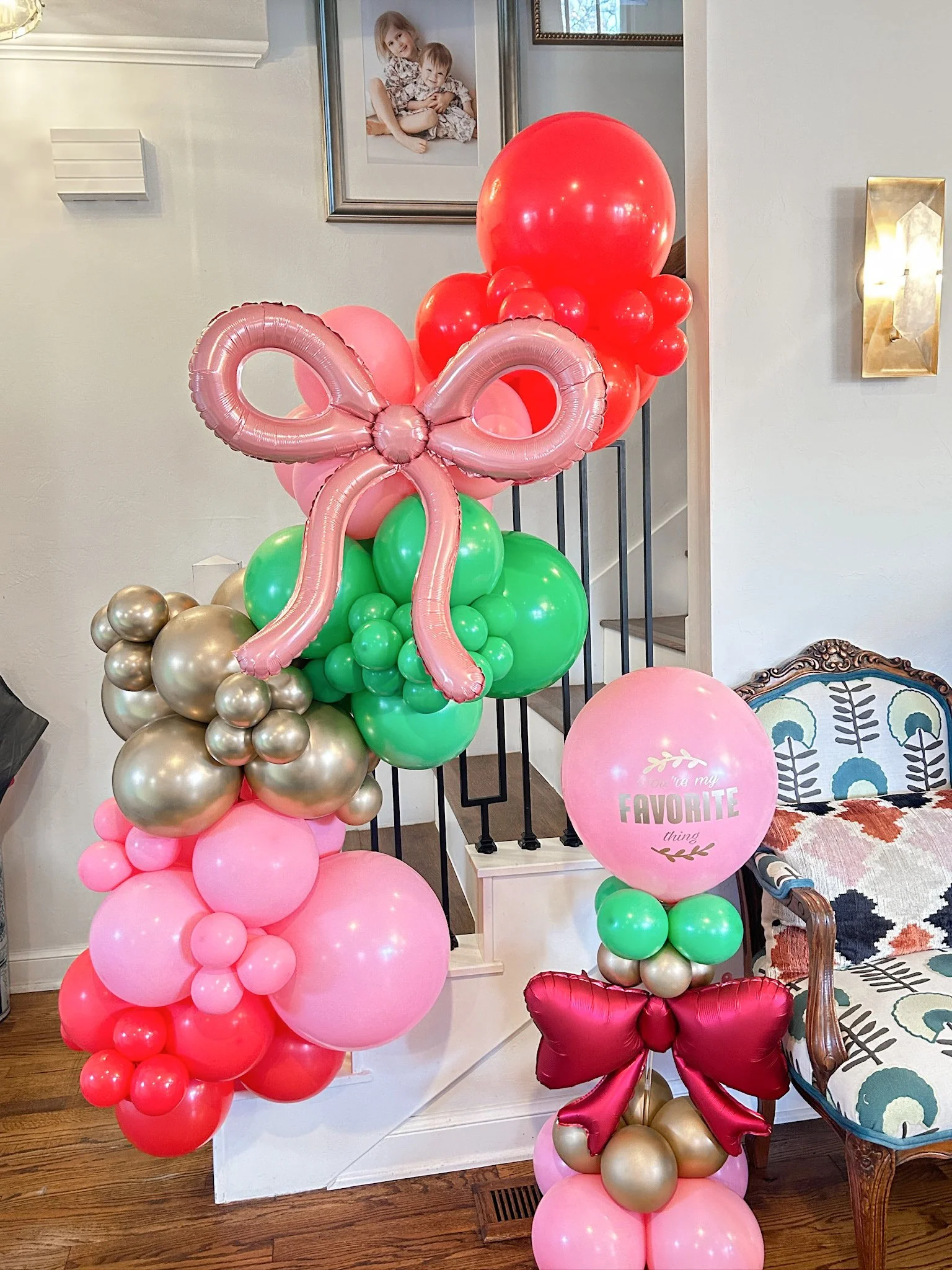 Colorful balloon arrangement with pink, red, green, gold, and rose gold balloons, including a large pink bow-shaped balloon and a pink balloon with the words "You are my favorite thing" written on it, inside a home near a staircase and a floral-patte