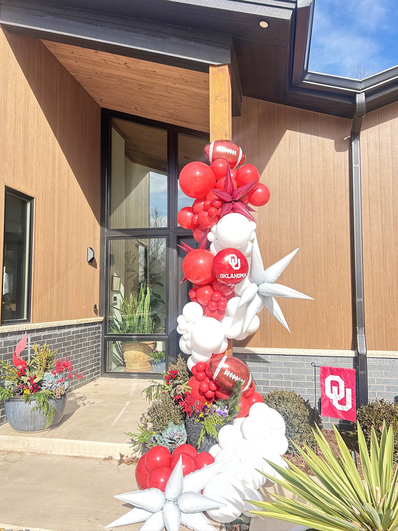 Decorative arrangement of red, white, and star-shaped balloons with Oklahoma Sooners logos, outside a building with a wooden exterior, plants, and a sign.