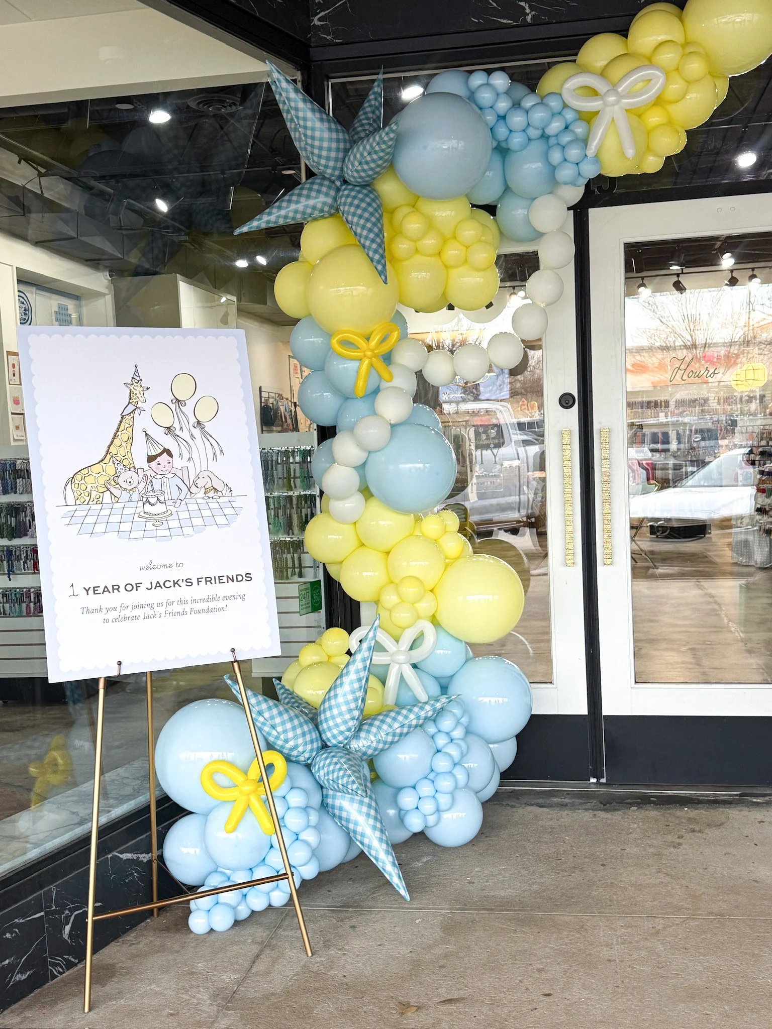 Balloon arrangement of yellow, blue, and white balloons with blue checked fabric pinwheels, set up outside a storefront. A sign indicates it celebrates one year of Jack's friends.