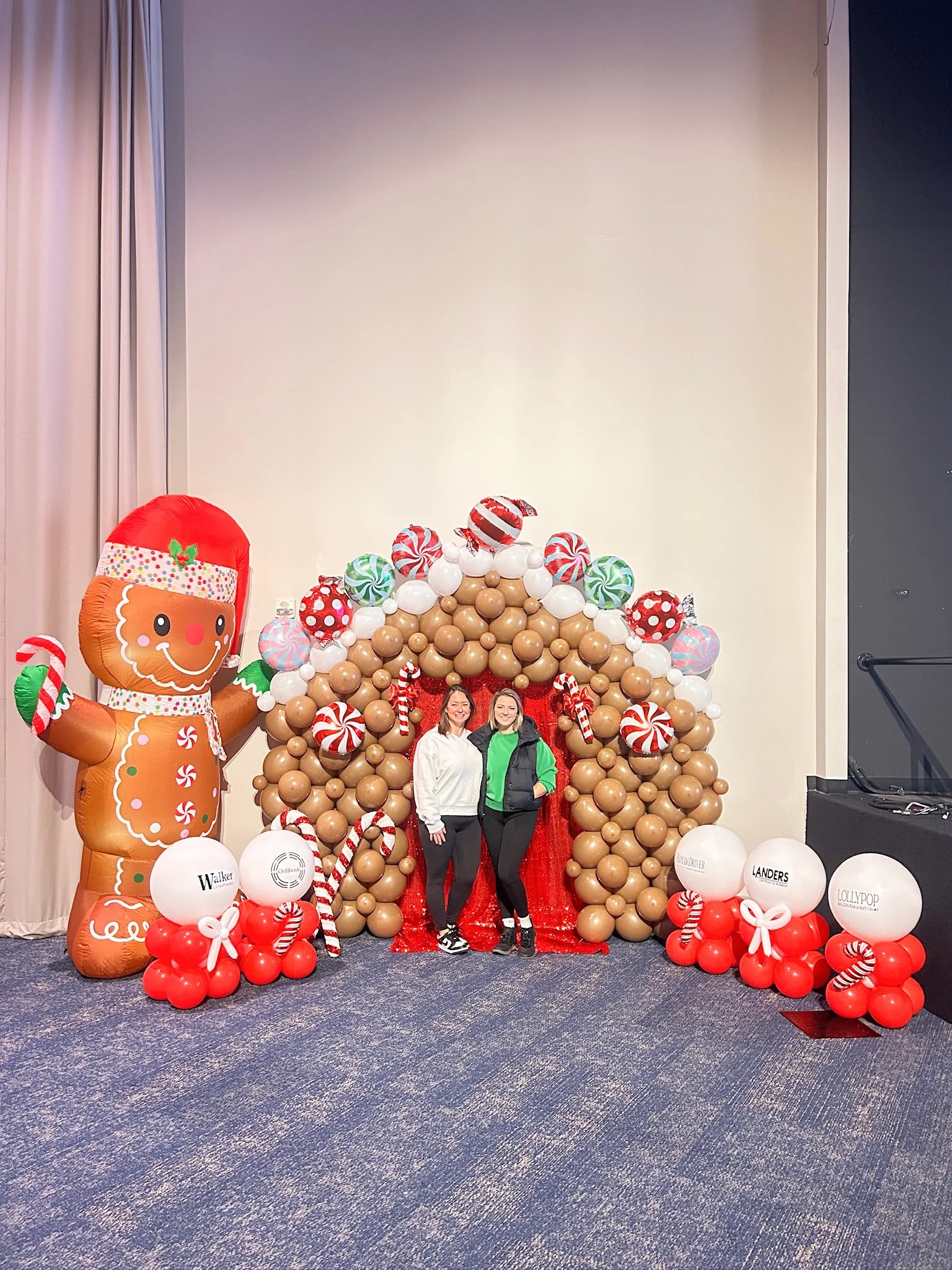 Two women standing in front of a holiday-themed balloon arch and a giant gingerbread man balloon