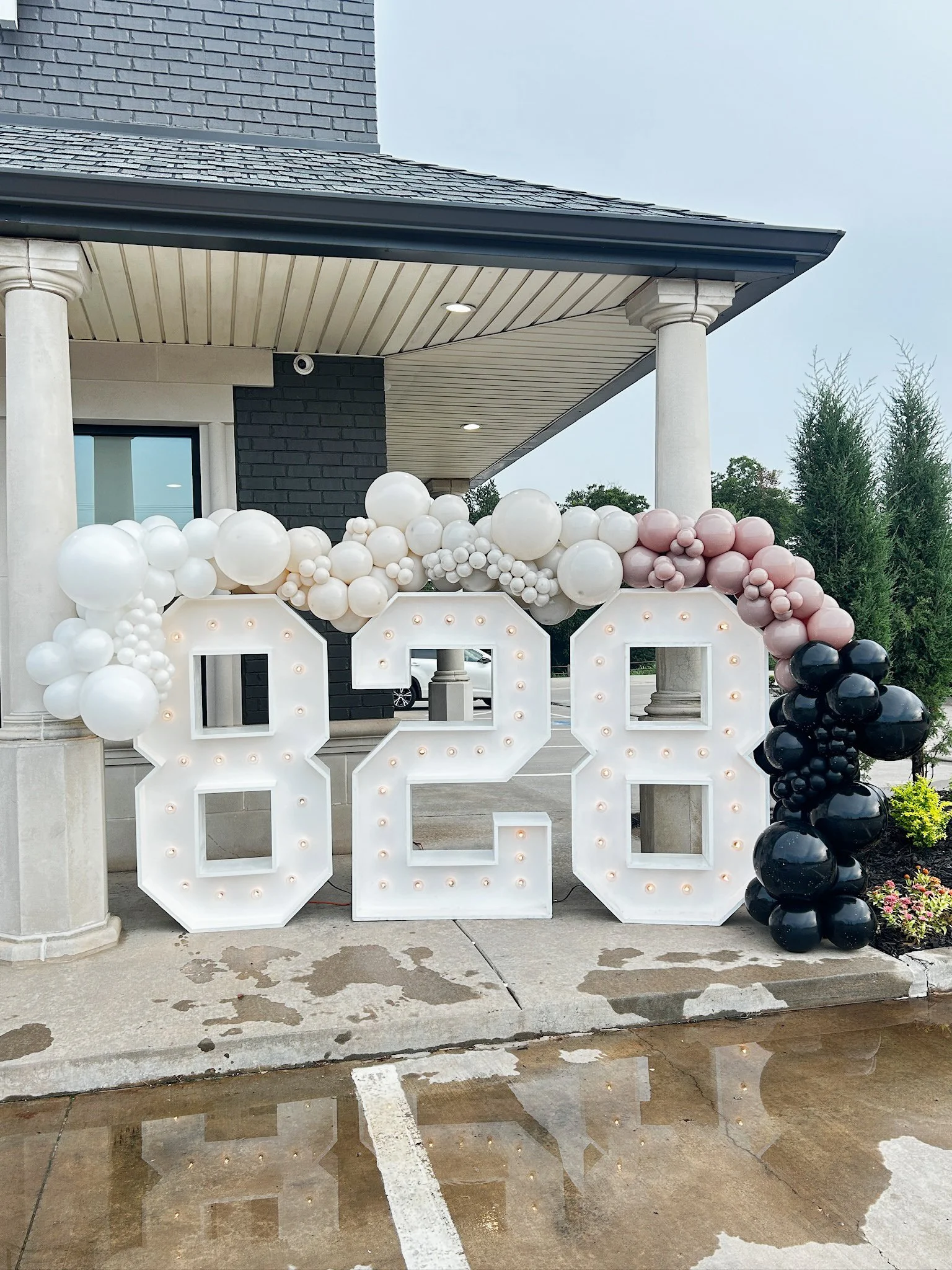 Large white illuminated numbers reading 2028, decorated with balloons in white, pink, and black, placed on a sidewalk outside a building with columns and a black and gray exterior.