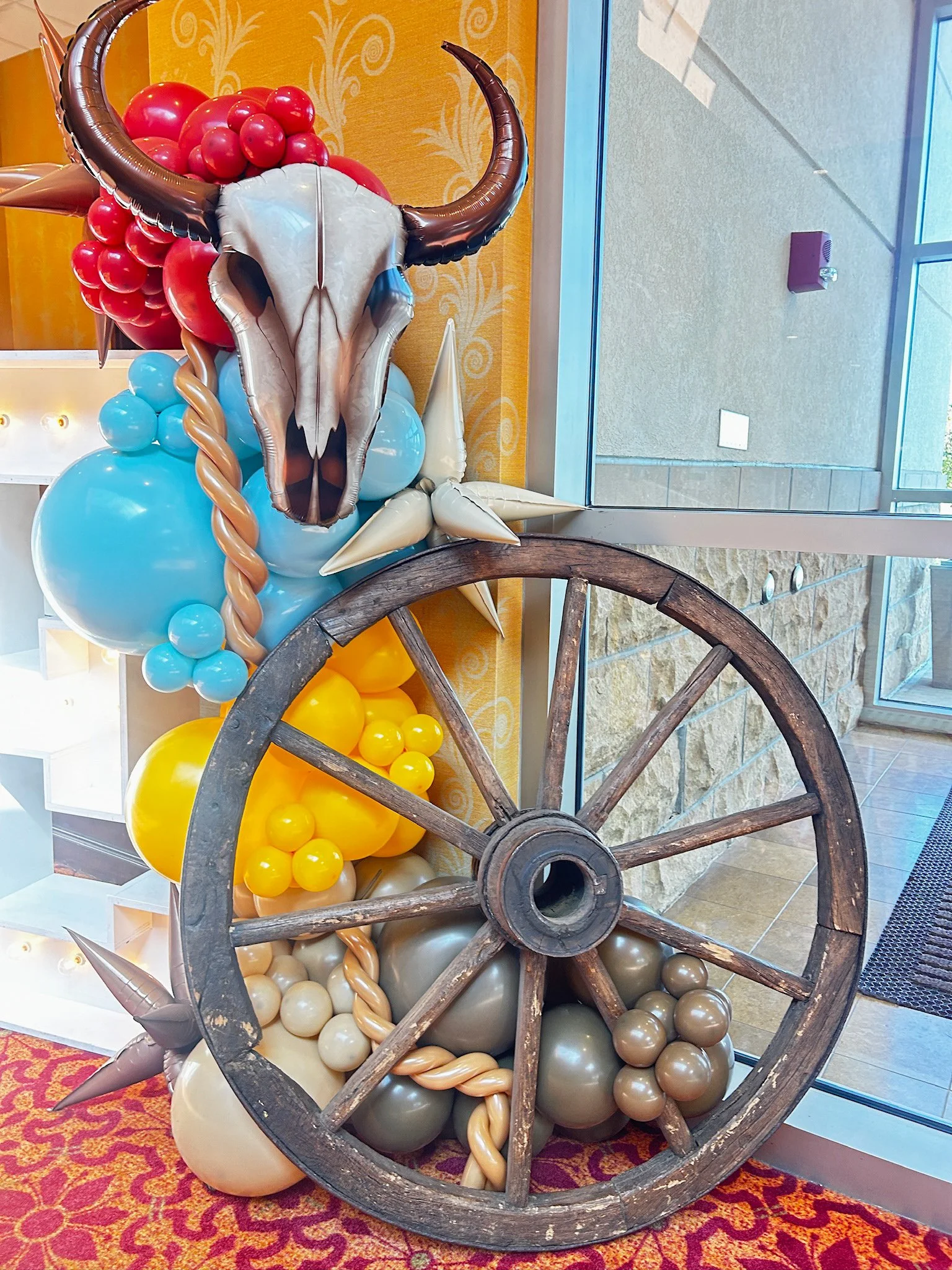 Decorative balloon sculpture featuring a buffalo skull with horns, surrounded by various colored balloons, a wooden wagon wheel, and a star-shaped ornament.
