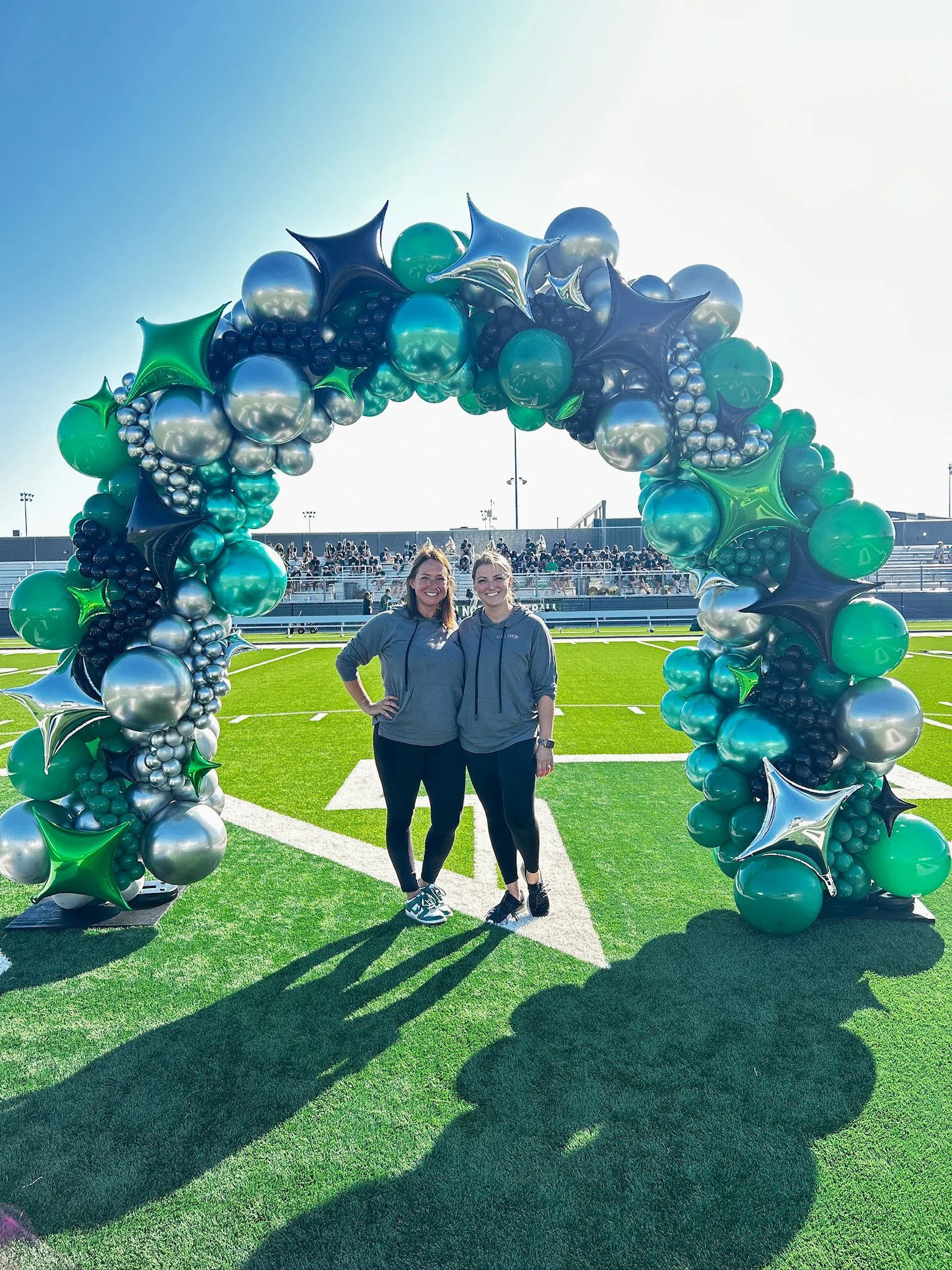 Two women standing under a colorful balloon arch on a sports field with bleachers and spectators in the background.