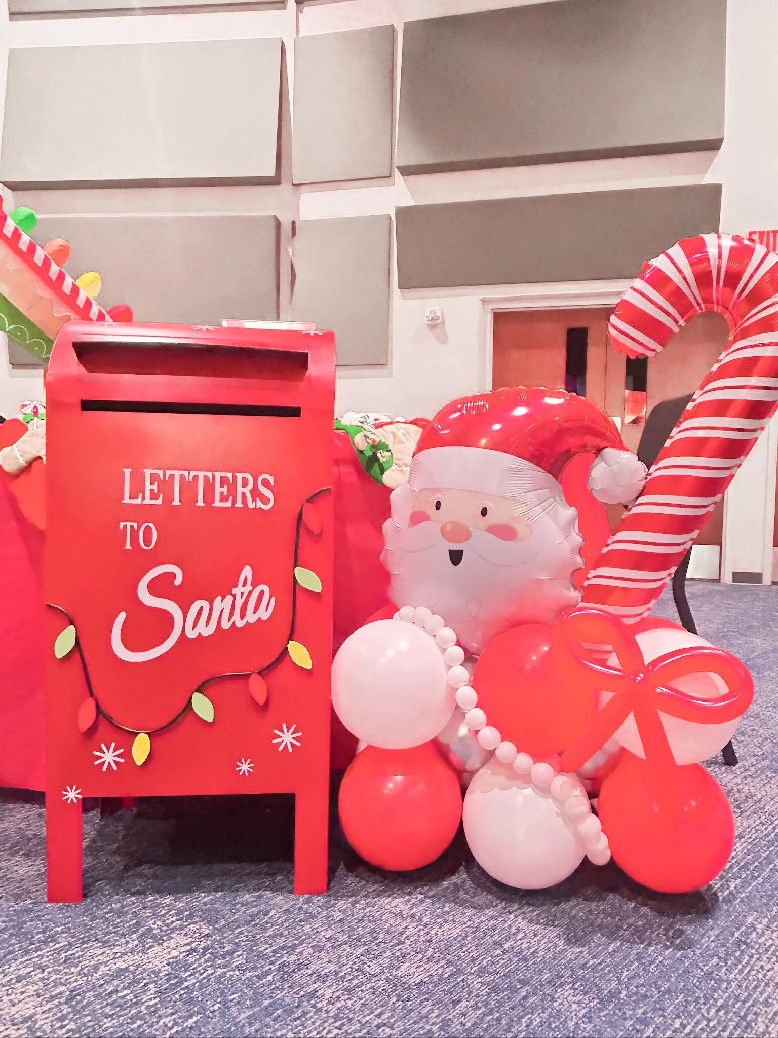 Christmas decorations including a red mailbox labeled 'Letters to Santa,' a Santa face balloon, and red and white balloons with a candy cane and bow, in a room with soundproof panels on the wall.