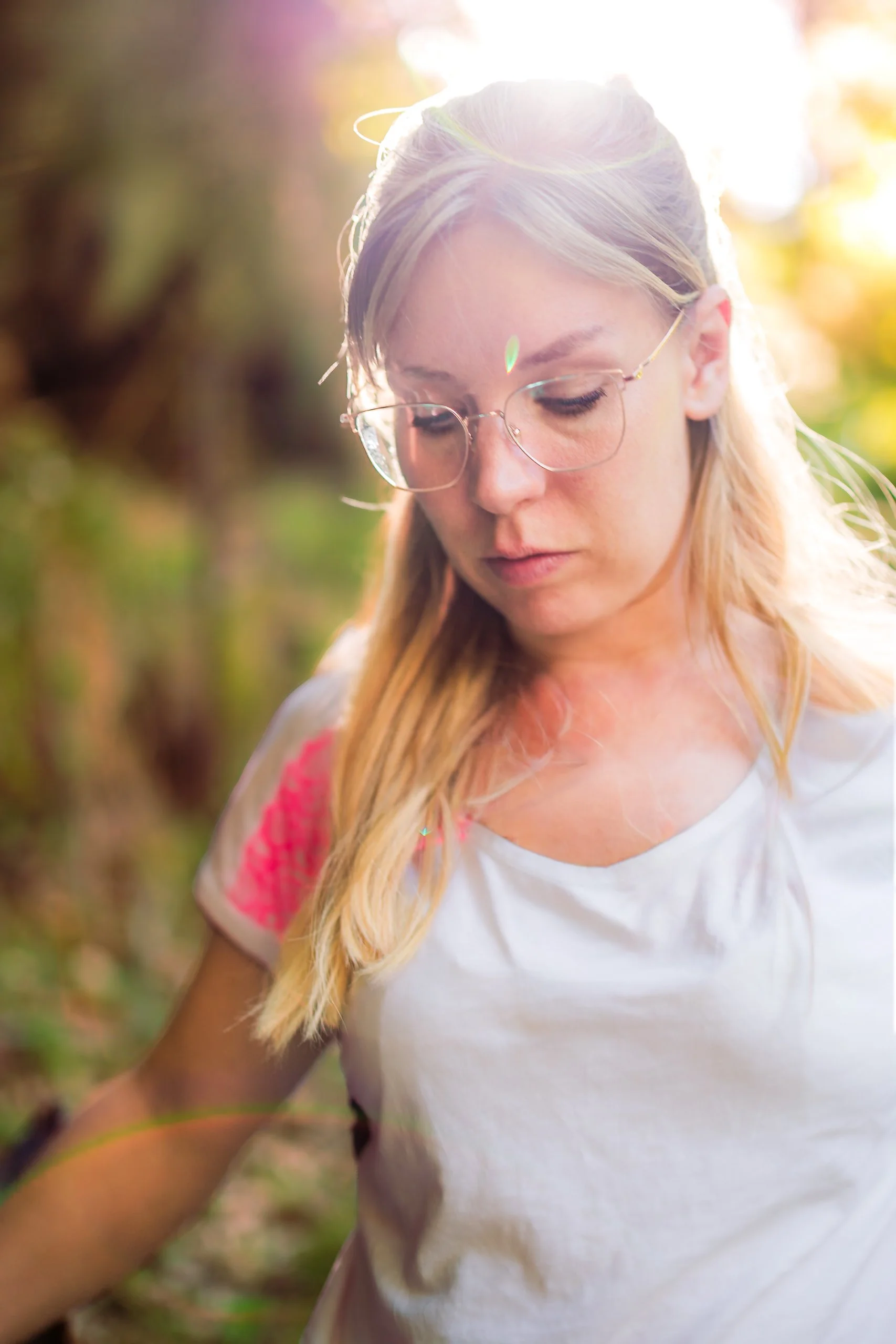Junge Frau mit bräunlichen Haaren, Gläsern und weißem T-Shirt, die draußen in der Natur bei Sonnenlicht steht.