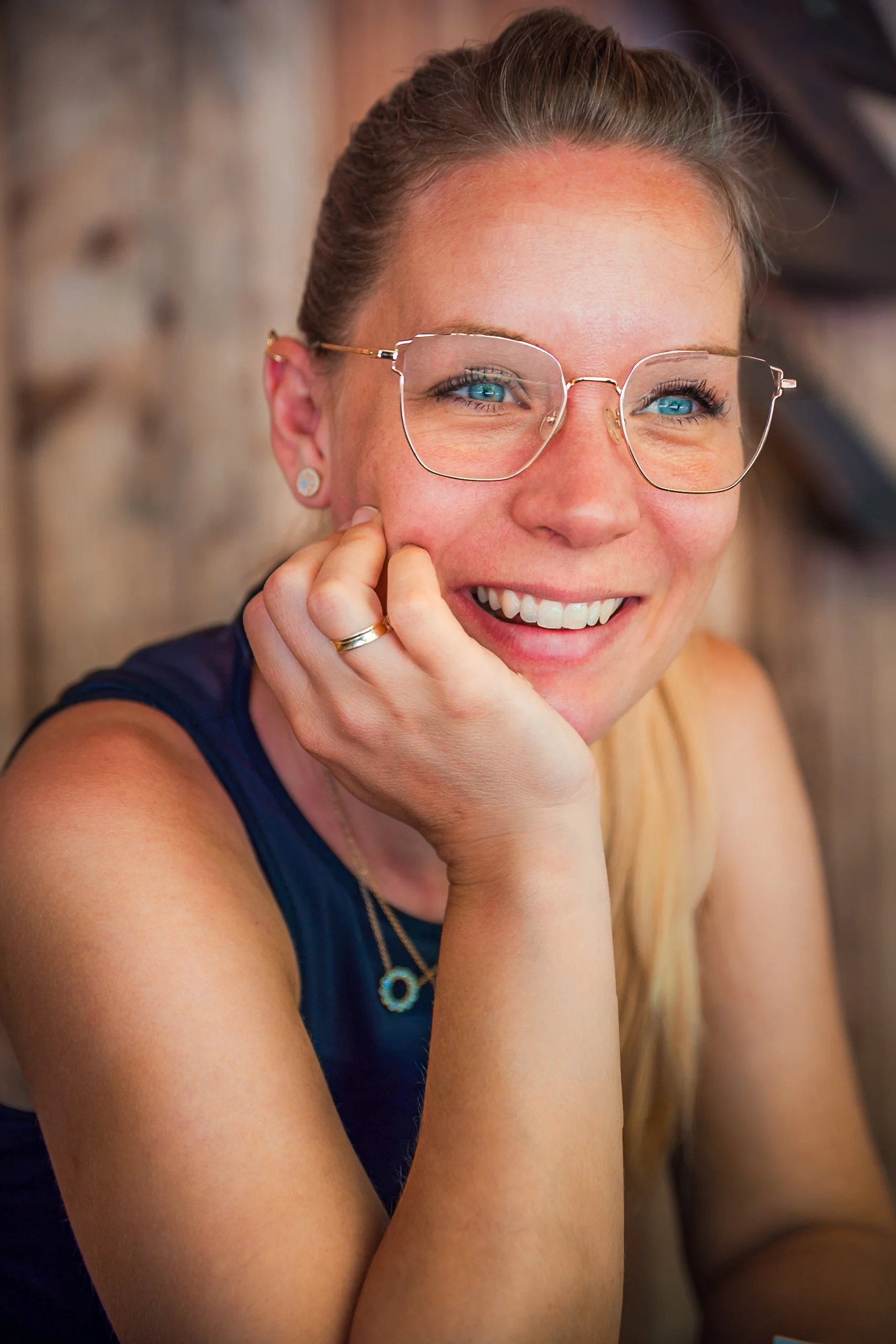 Nahaufnahme einer lachenden Frau mit blonden Haaren, Brille, Ohrringen und einem Halsketten, sitzend in einem rustikalen Holzraum.
