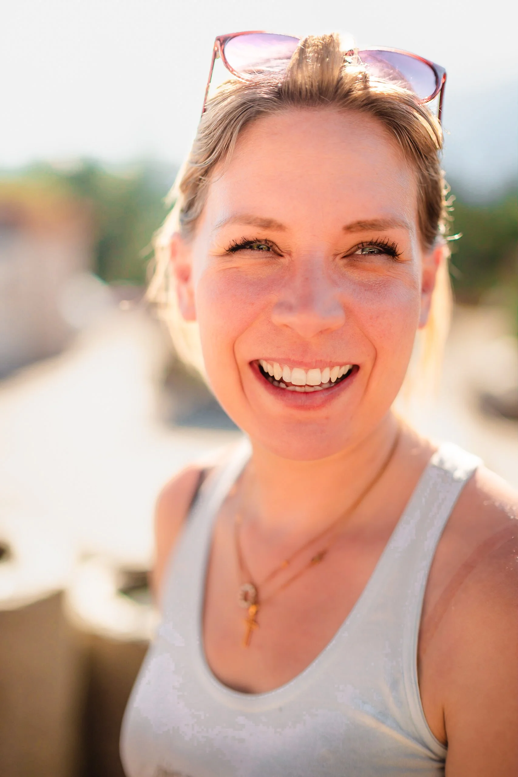 Junge Frau mit Sonnenbrille auf dem Kopf lächelt breit in die Kamera, im Sonnenlicht.