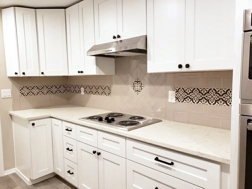White kitchen cabinets with black knobs, stainless steel range hood, beige tiled backsplash with decorative black and brown tile accents, white countertop, and a built-in cooktop with four burners.