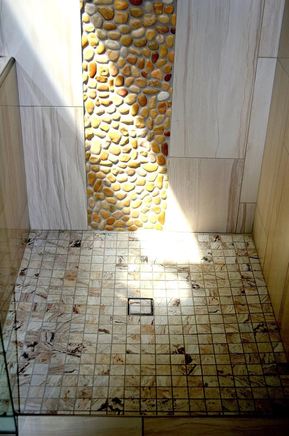 Inside a shower area with a mosaic pebble wall and beige marble tiles on the walls and floor, featuring a square drain at the floor's center.