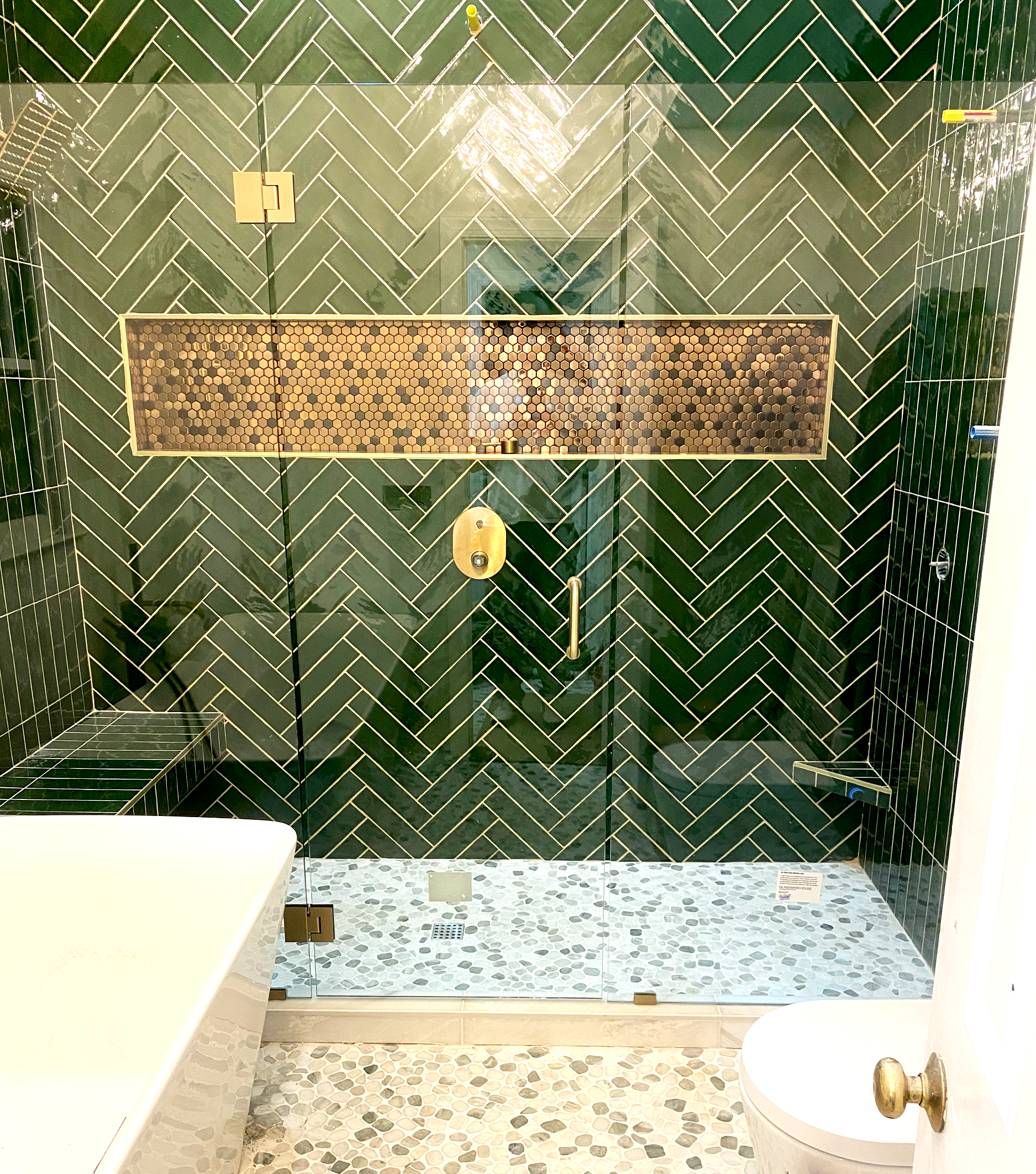 Bathroom with a glass shower enclosure, green herringbone tile on the walls, and a floating bench. The shower features a horizontal band of small dark hexagonal tiles. The floor of the shower has pebble tiling, and there is a gold showerhead and hand