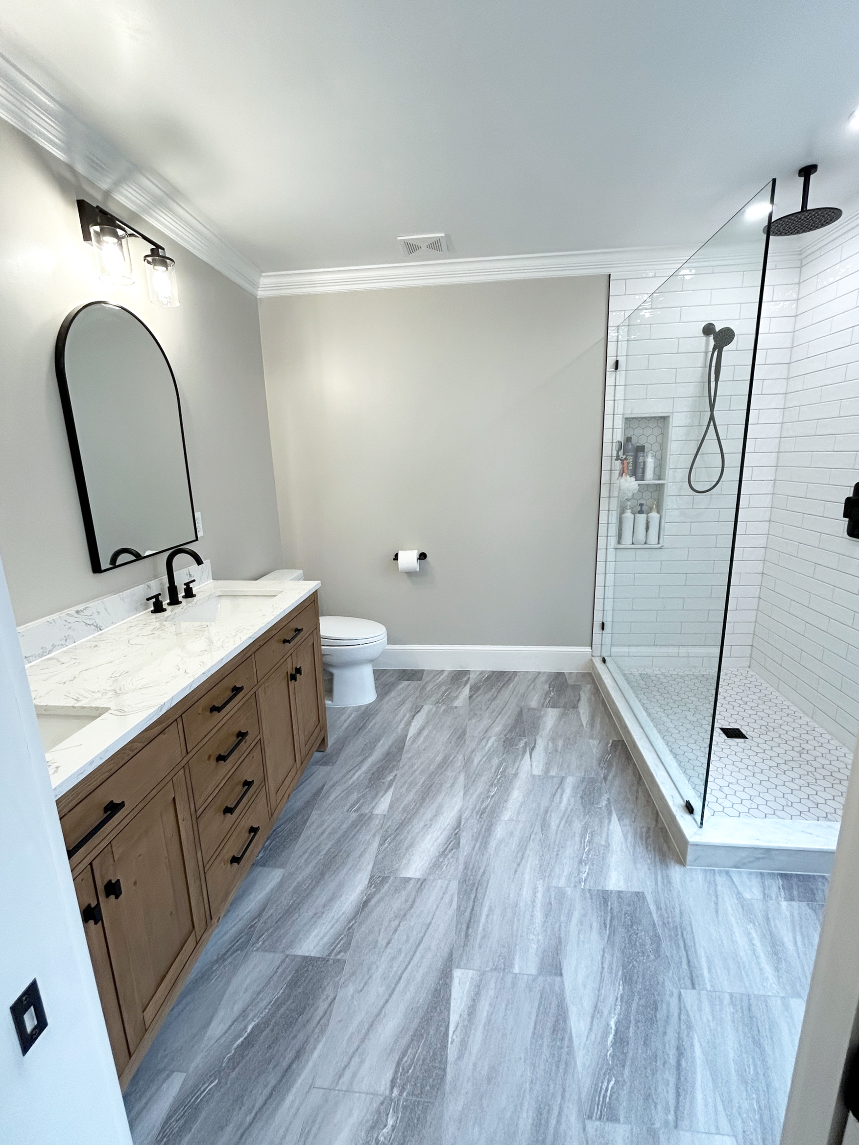 Modern bathroom with a large walk-in shower, wooden vanity with marble countertop, and a toilet, featuring light gray walls and wooden floor tiles.